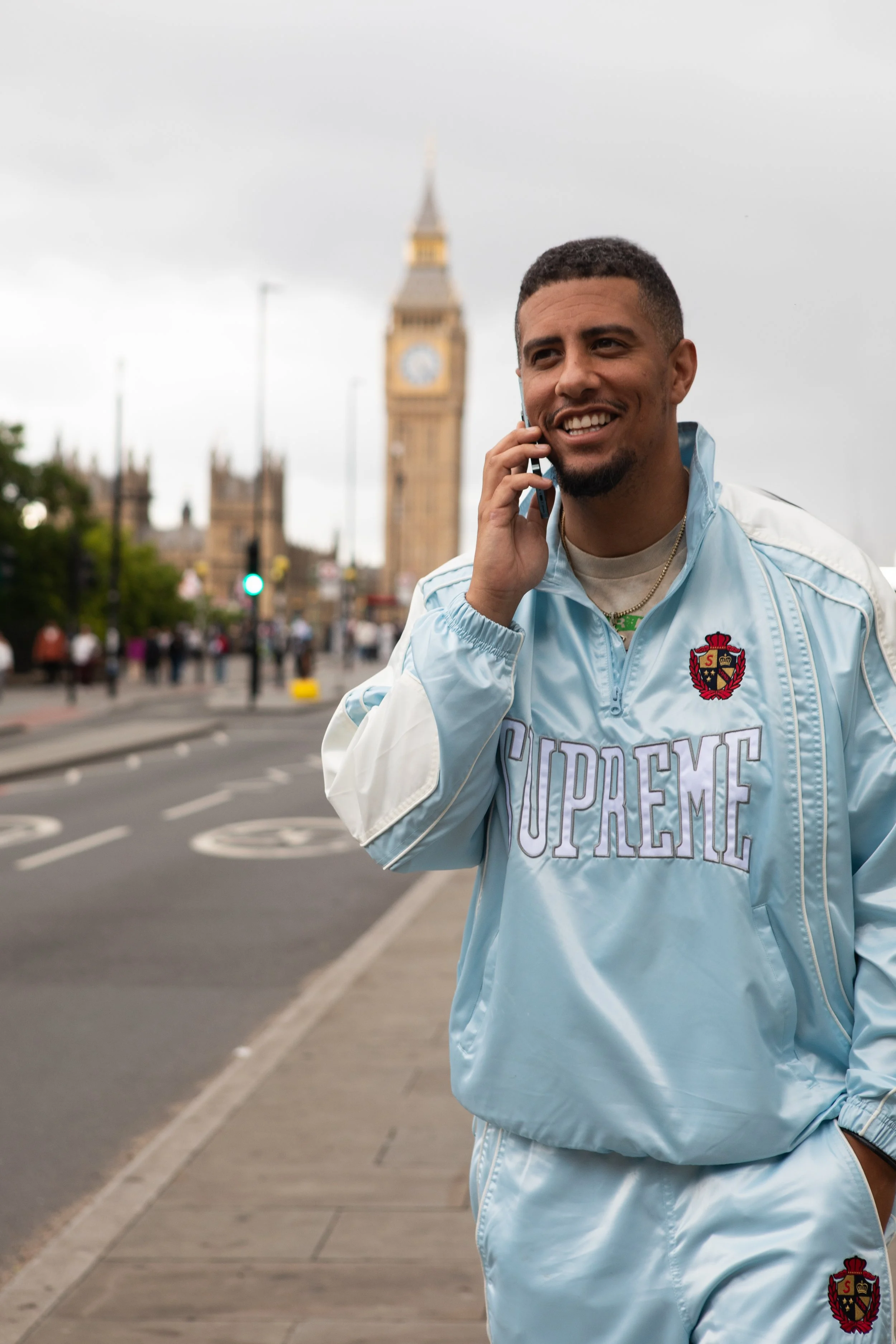 A man, Jake Amankwaah  in a light blue Supreme jacket is smiling and talking on his phone on a city sidewalk with Big Ben in the background.