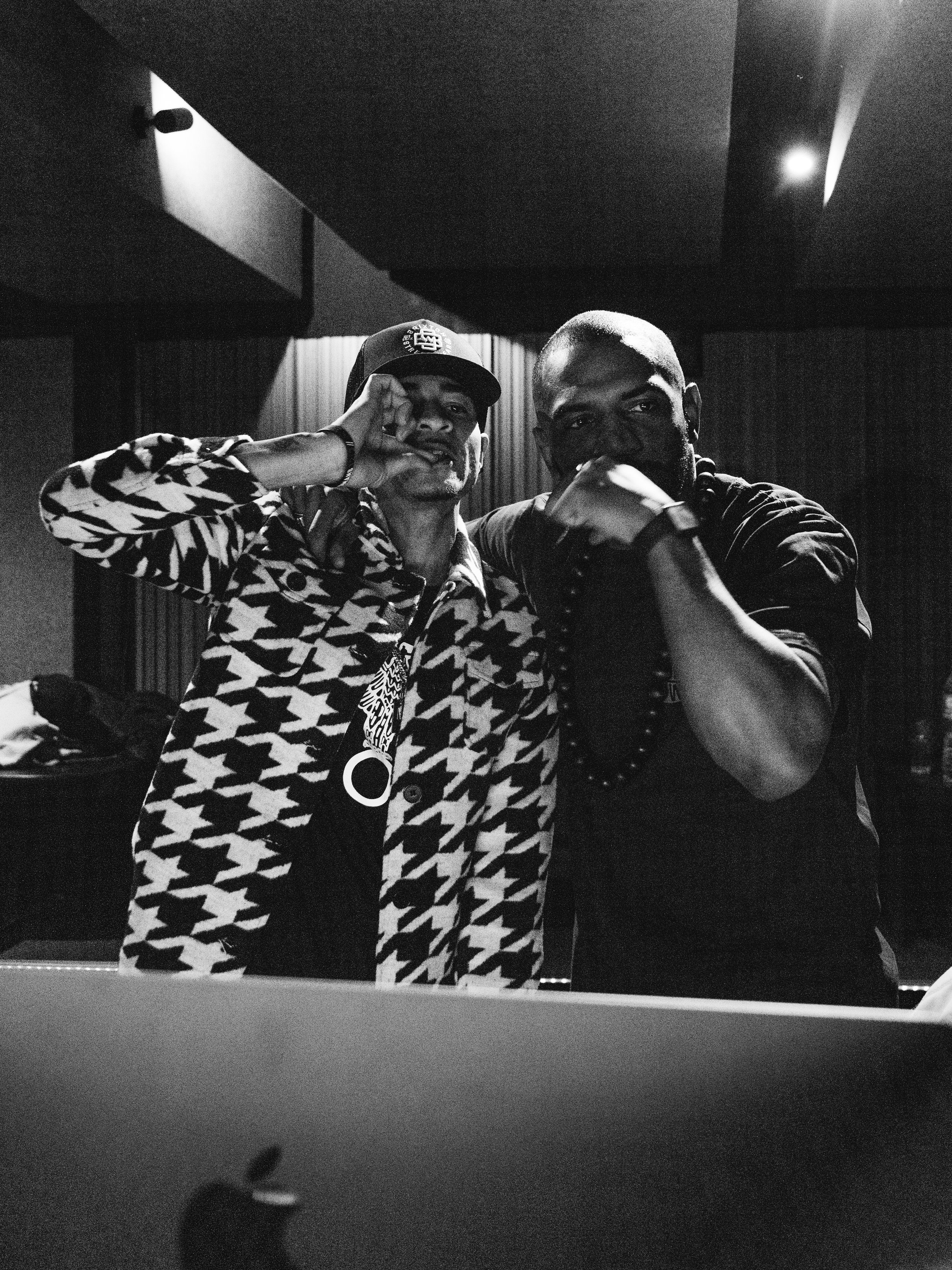 Two men pose for a black and white photo in a recording studio, with one wearing a patterned jacket and hat, and the other in dark clothing with accessories, both making hand gestures.