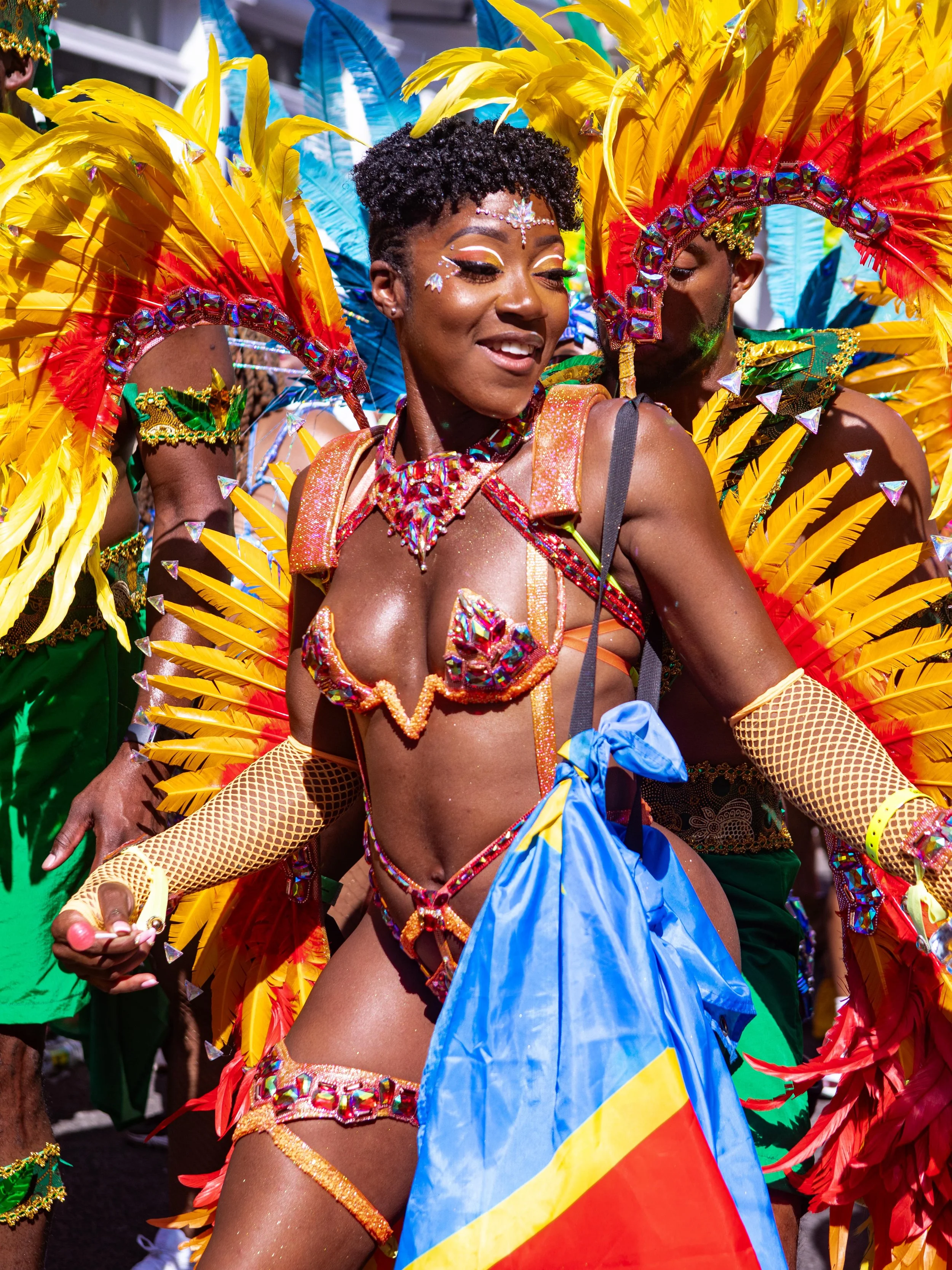 A woman in a vibrant Carnival costume with yellow, red, and blue feathers, beaded jewelry, and glitter makeup, holding a flag with blue and yellow stripes.