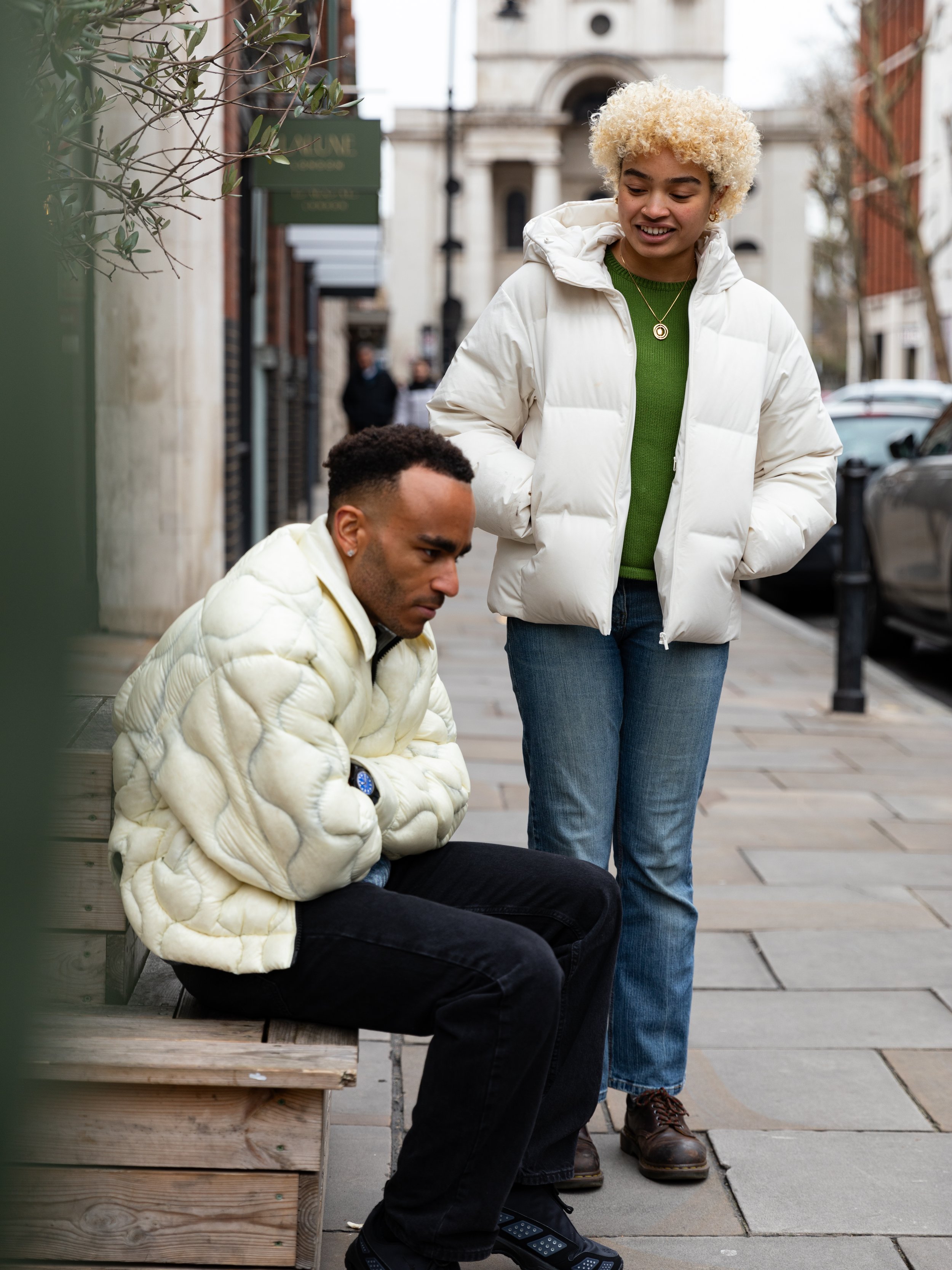 A woman standing and looking down at a man sitting on a bench on a city sidewalk. Both are wearing white puffer jackets. The woman has curly blonde hair, and the man has short dark hair. Behind them is an urban street scene with buildings and parked 