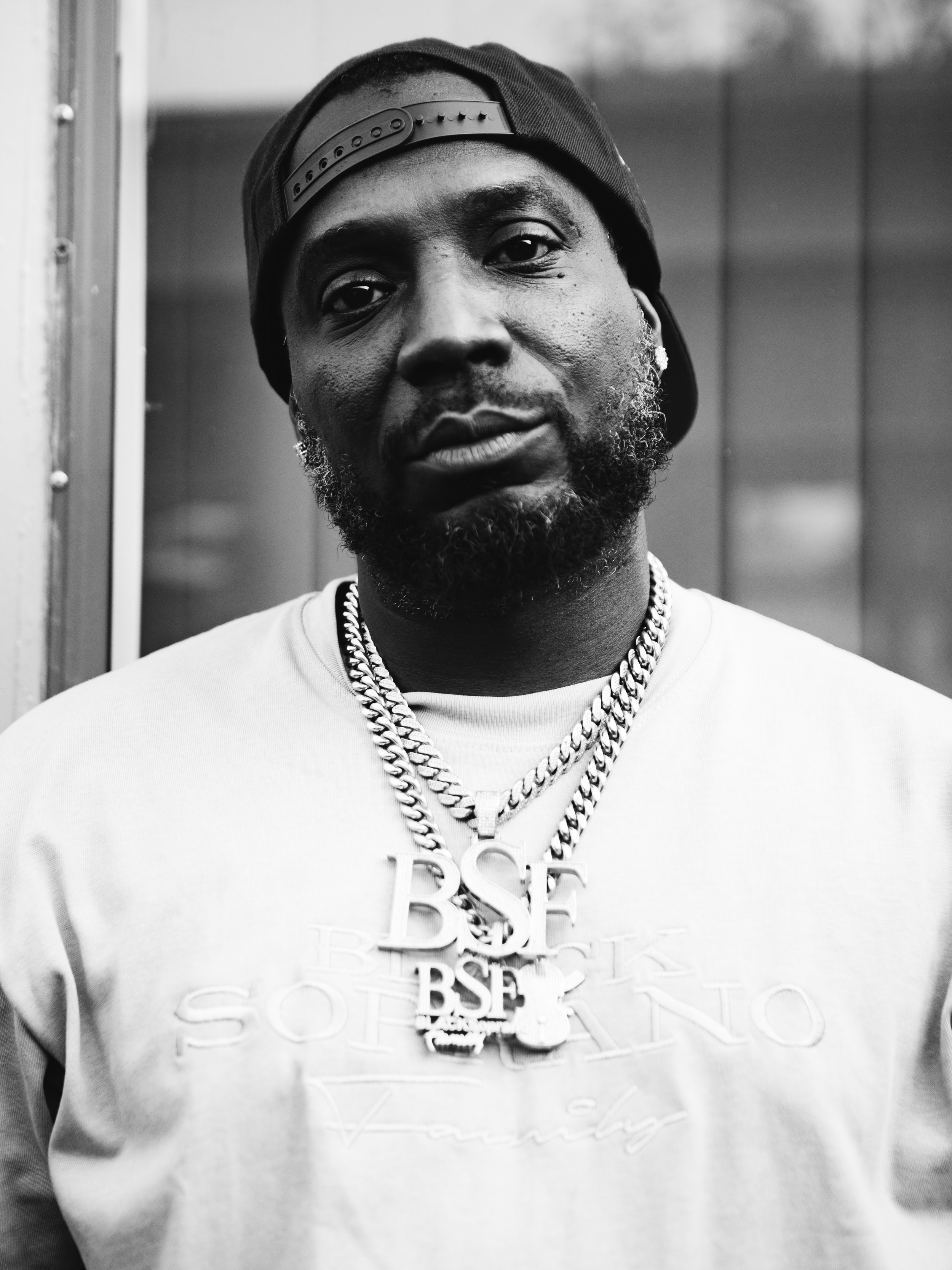 A Black man, B$F City Boy,  with a beard wearing a backwards baseball cap, a light-colored sweatshirt, and multiple chains around his neck, including one with the letters "B$F." Backstage at Jazz Cafe Festival, London