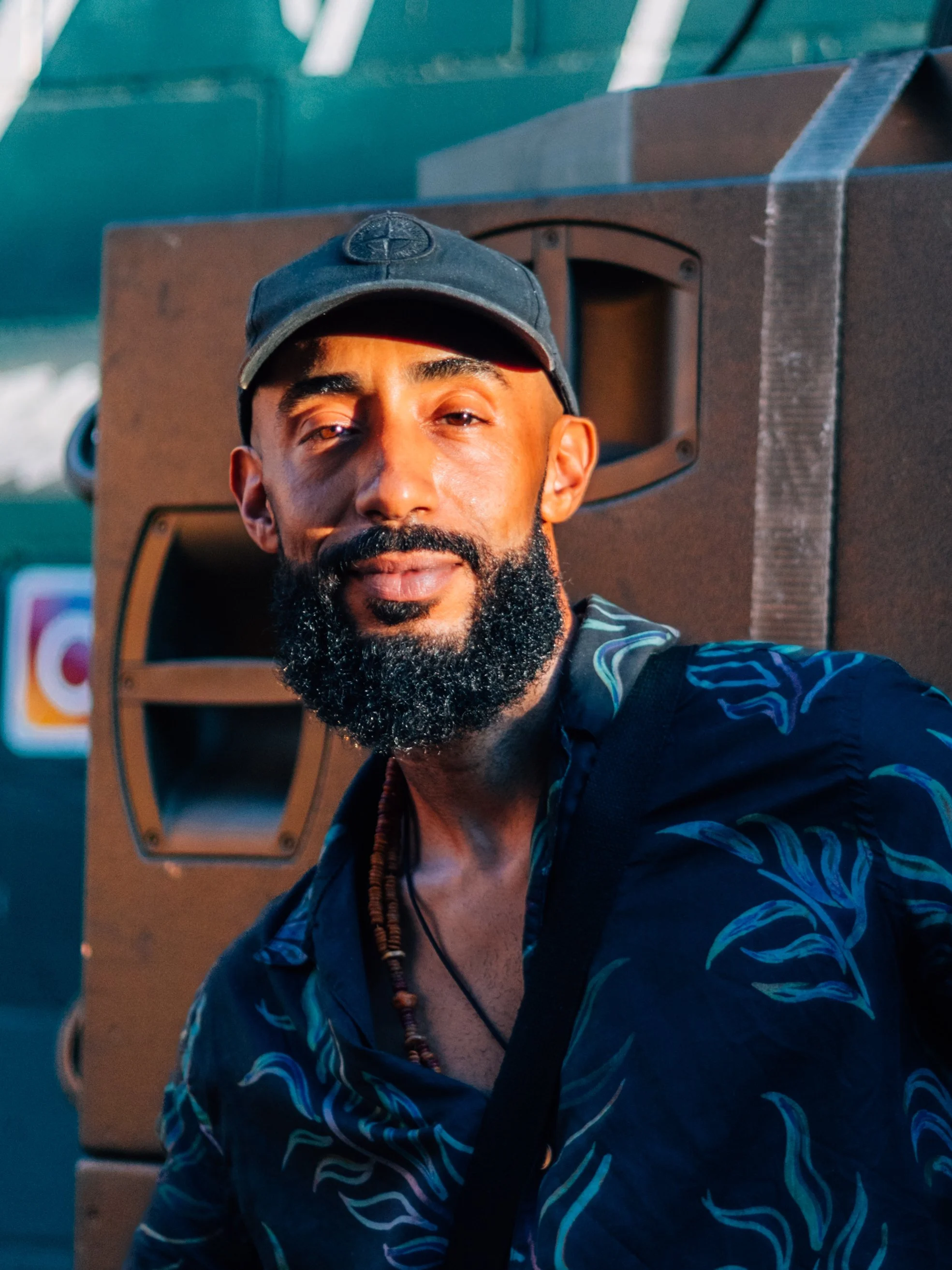 A man with a beard and mustache wearing a dark cap and a dark patterned shirt, standing outdoors near a large speaker, with a smile and relaxed expression. Hackney Reggae Festival, London in the sun
