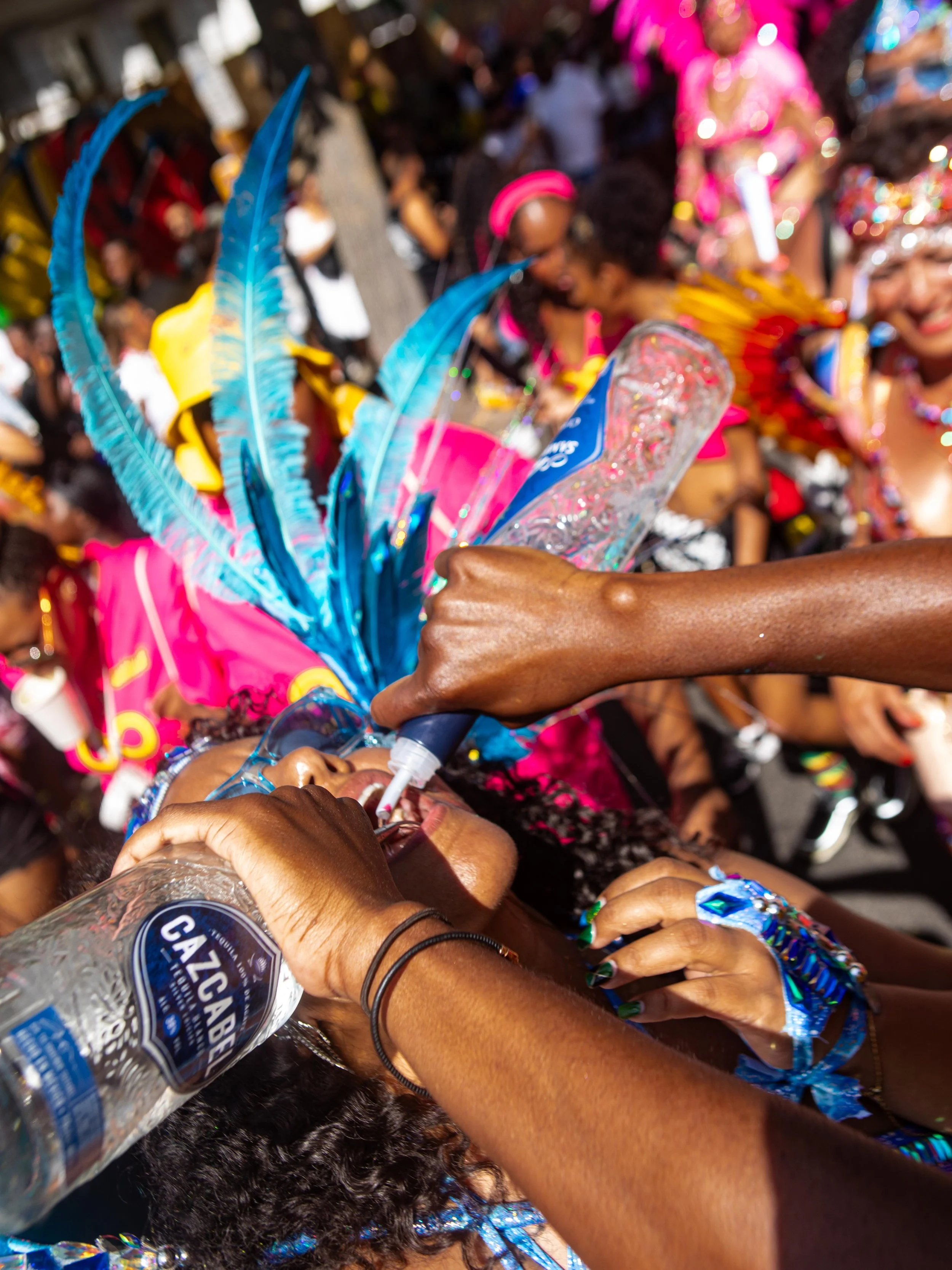A person is drinking Tequila from a bottle during a colorful outdoor festival or parade, Notting Hill Carnival, surrounded by people dressed in vibrant costumes and feathered accessories.