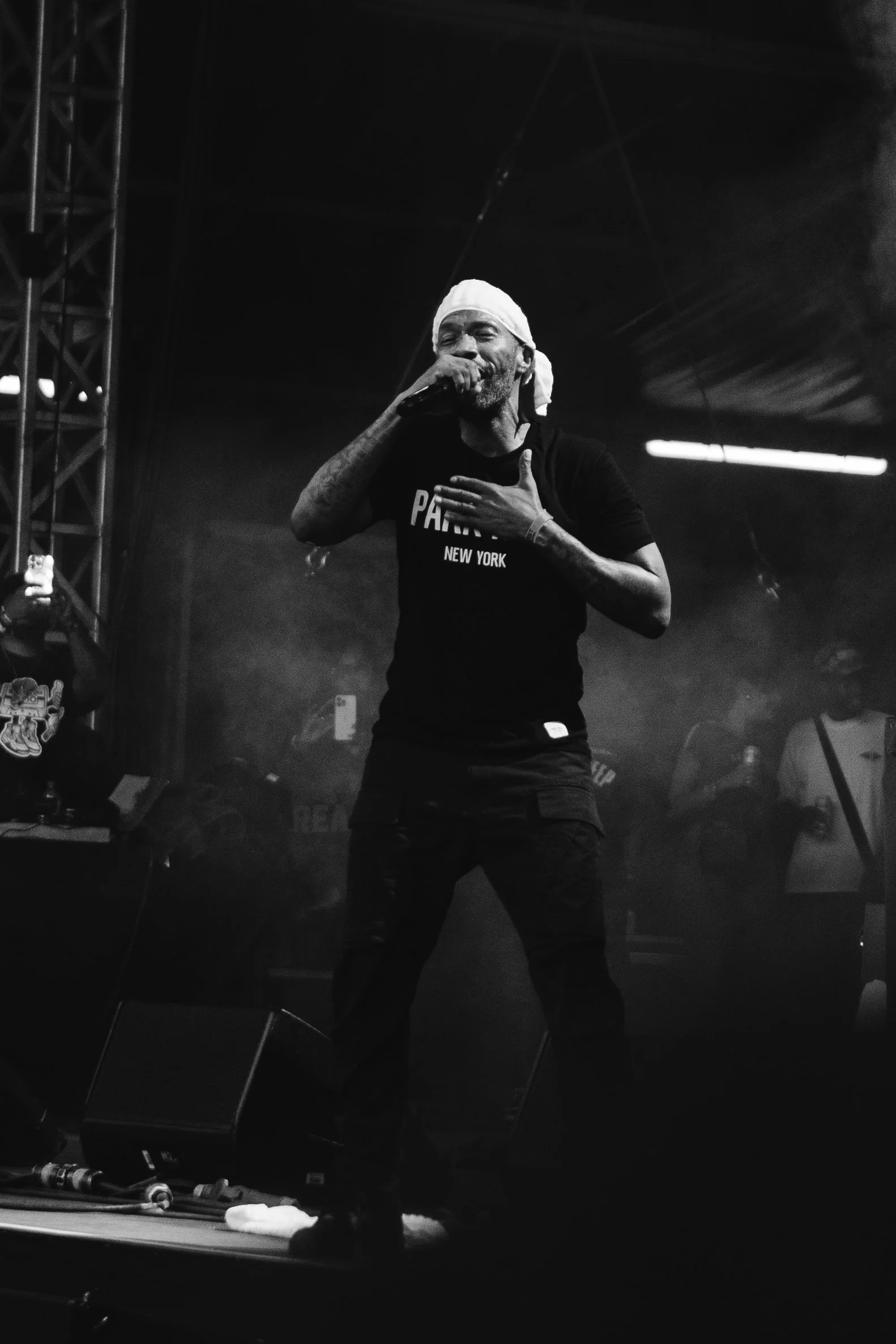 A male, Redman, performer on stage singing into a microphone, wearing a white bandana and black T-shirt, with a DJ setup and other people in the background at South Facing Festival in Crystal Palace.