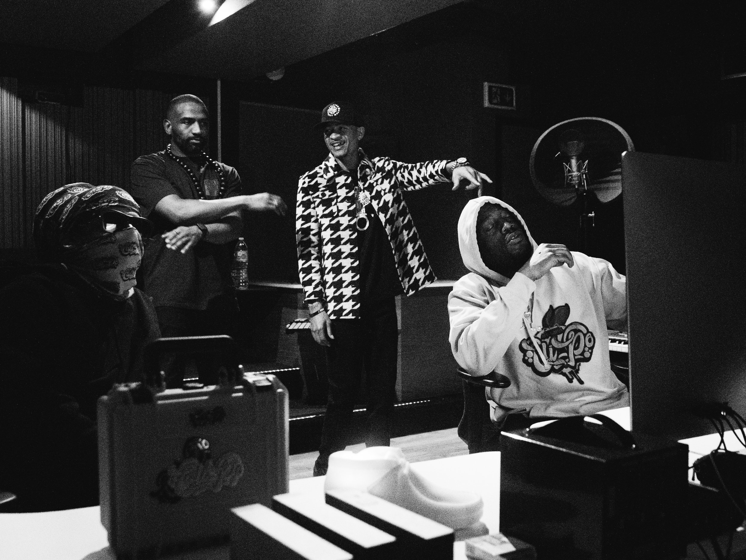 Four men in a recording studio; one seated at a computer wearing a hoodie and white cap, three standing behind, one with arm raised and wearing a patterned jacket, another with arms crossed and wearing a cap, and the third with a mask and helmet.