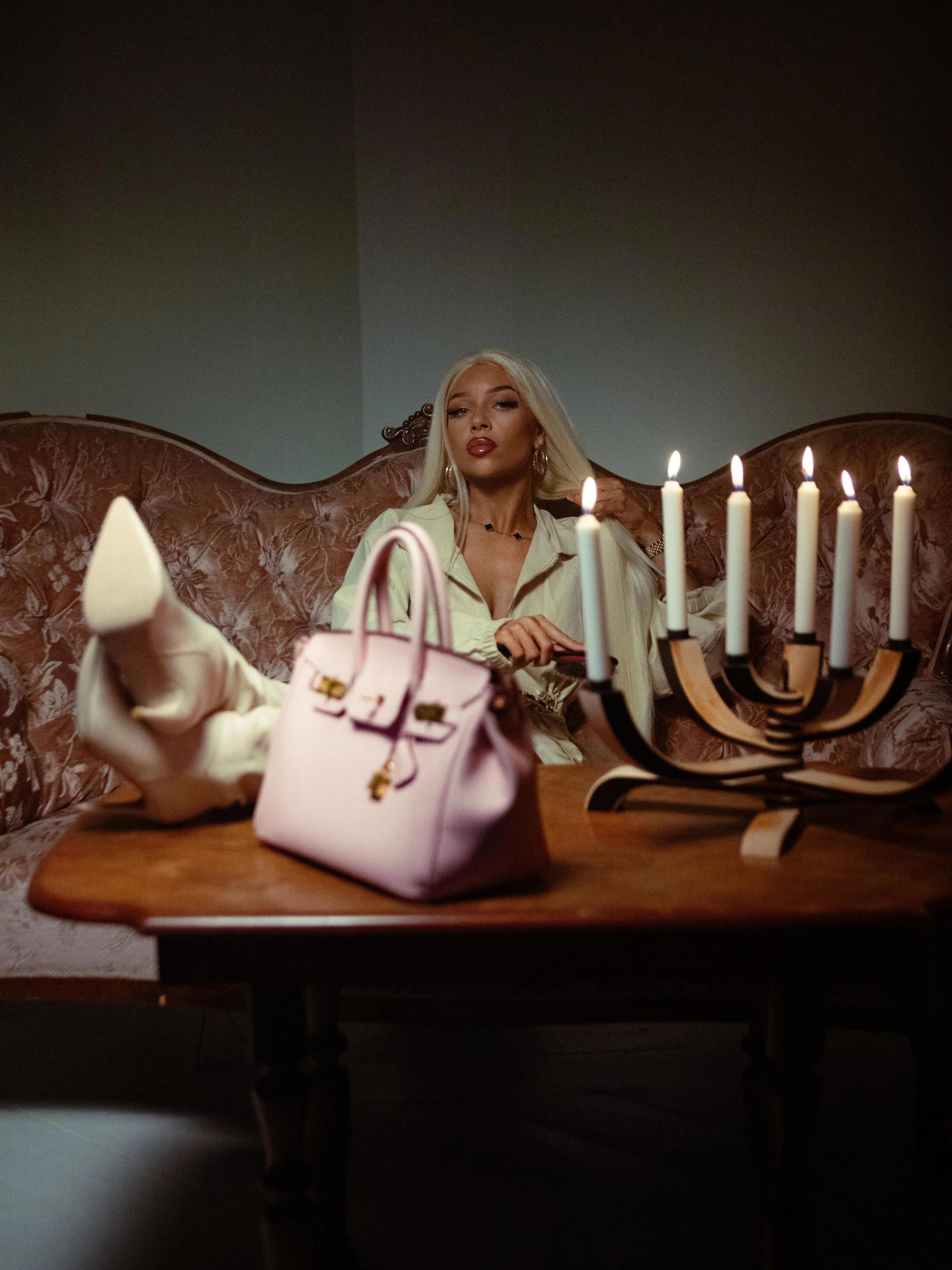 A woman with long blonde hair and gold hoops sitting on a vintage sofa, with a pink handbag on the table and a candelabrum with lit white candles in front of her.
