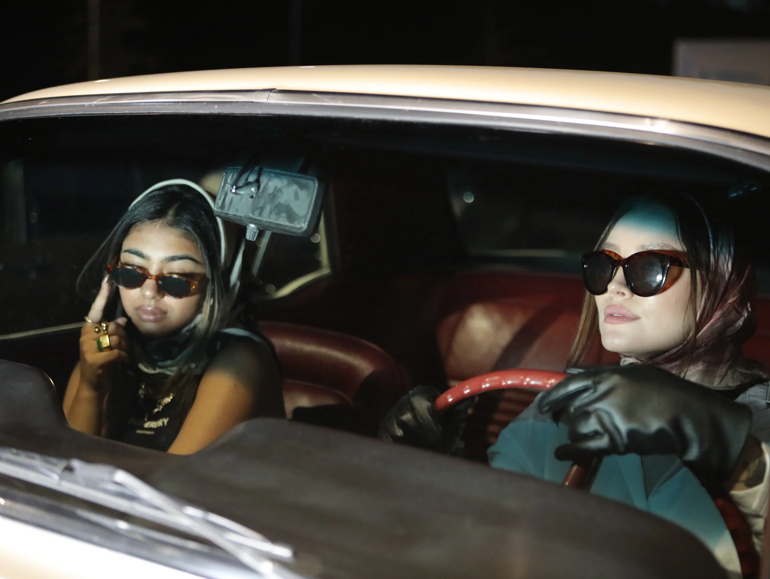 Two women sitting in a vintage car at night, both wearing sunglasses and dark clothing. The woman on the left has dark hair with highlights, wearing large sunglasses, and touching her face. The woman on the right, who is driving, has dark hair and is
