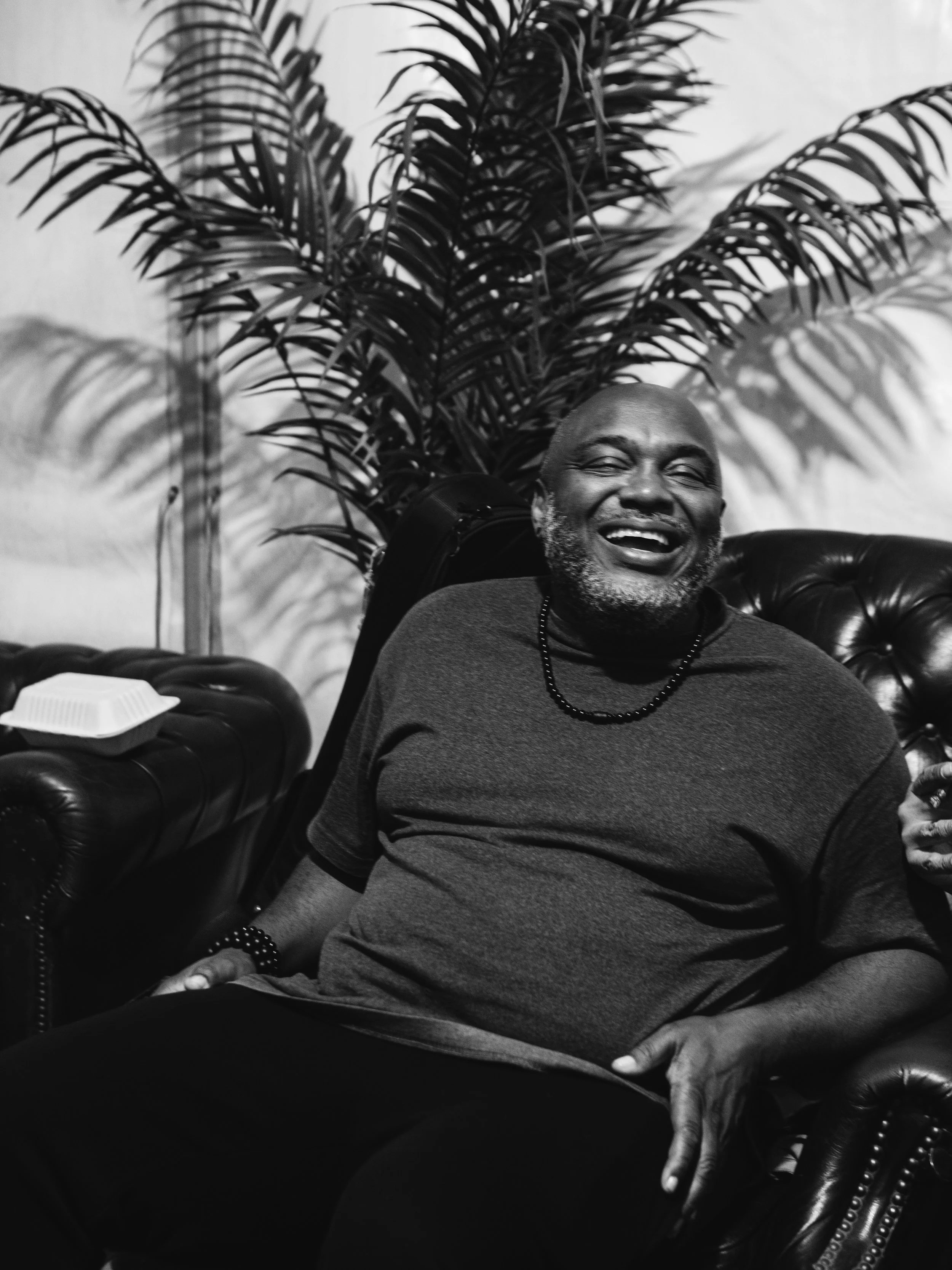 A joyful man with a beard sitting on a leather sofa, laughing, with a large potted plant behind him and a takeout food container on the sofa armrest. Nigel, Keyboardist from Rough Cut Band backstage at Boom Town Fair Sean Paul