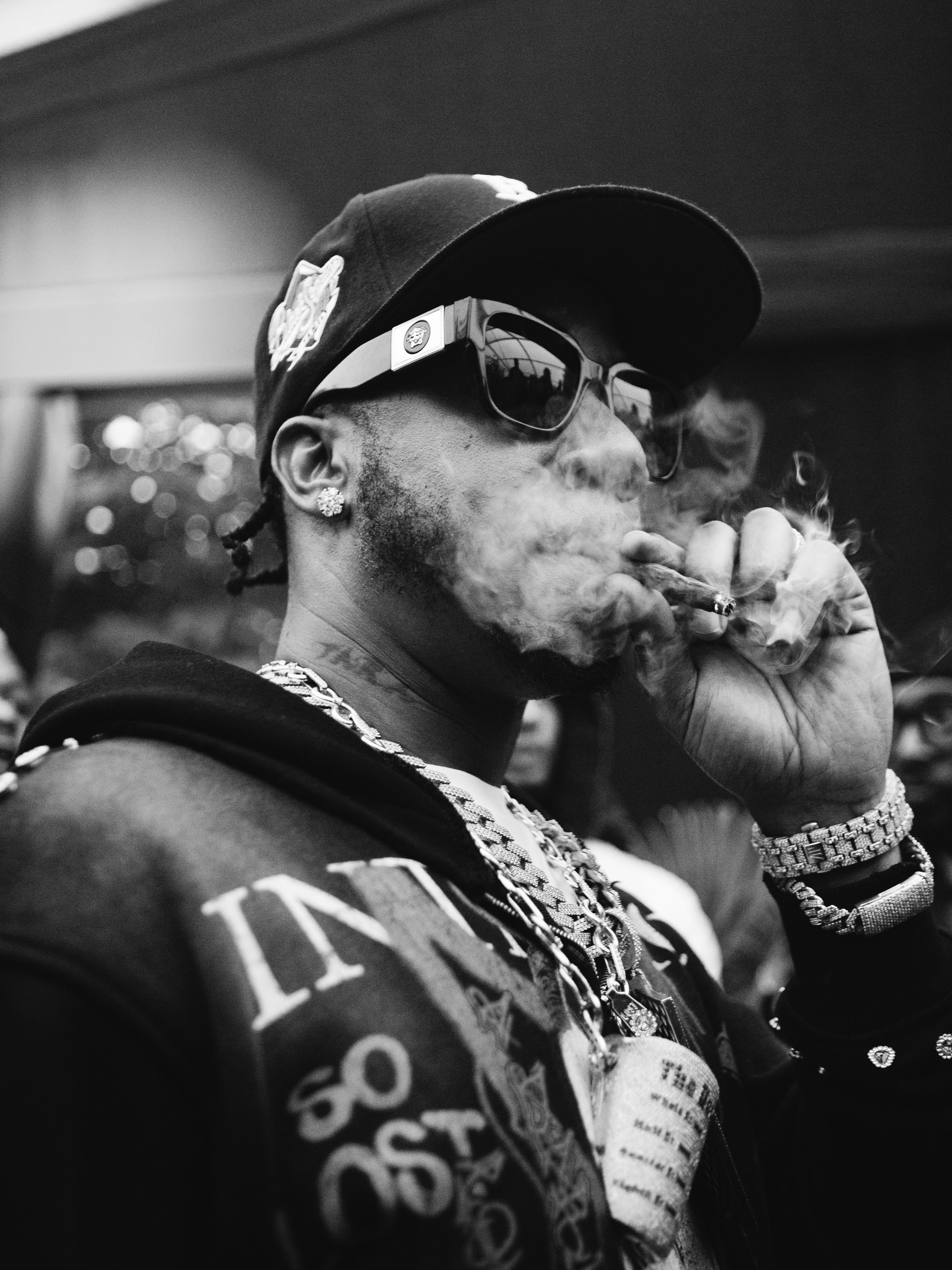 A man wearing sunglasses, a cap, and jewelry, smoking a cigar or cigarette, with visible smoke around him.