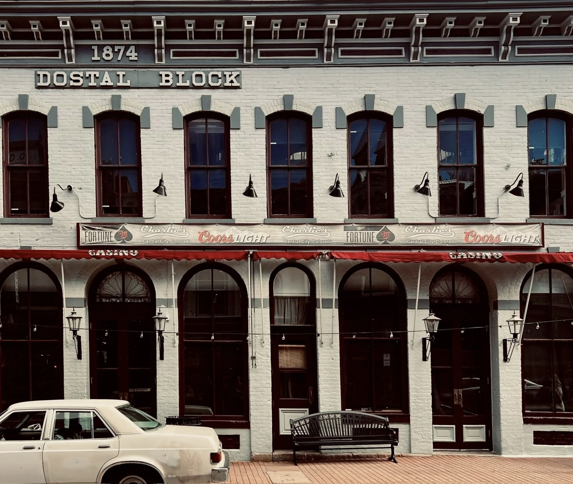 Street scene featuring a multi-story building with white brick walls, multiple arched windows, and attached exterior lights. A vintage car is parked in the foreground, and there is an outdoor bench and string lights along the sidewalk.