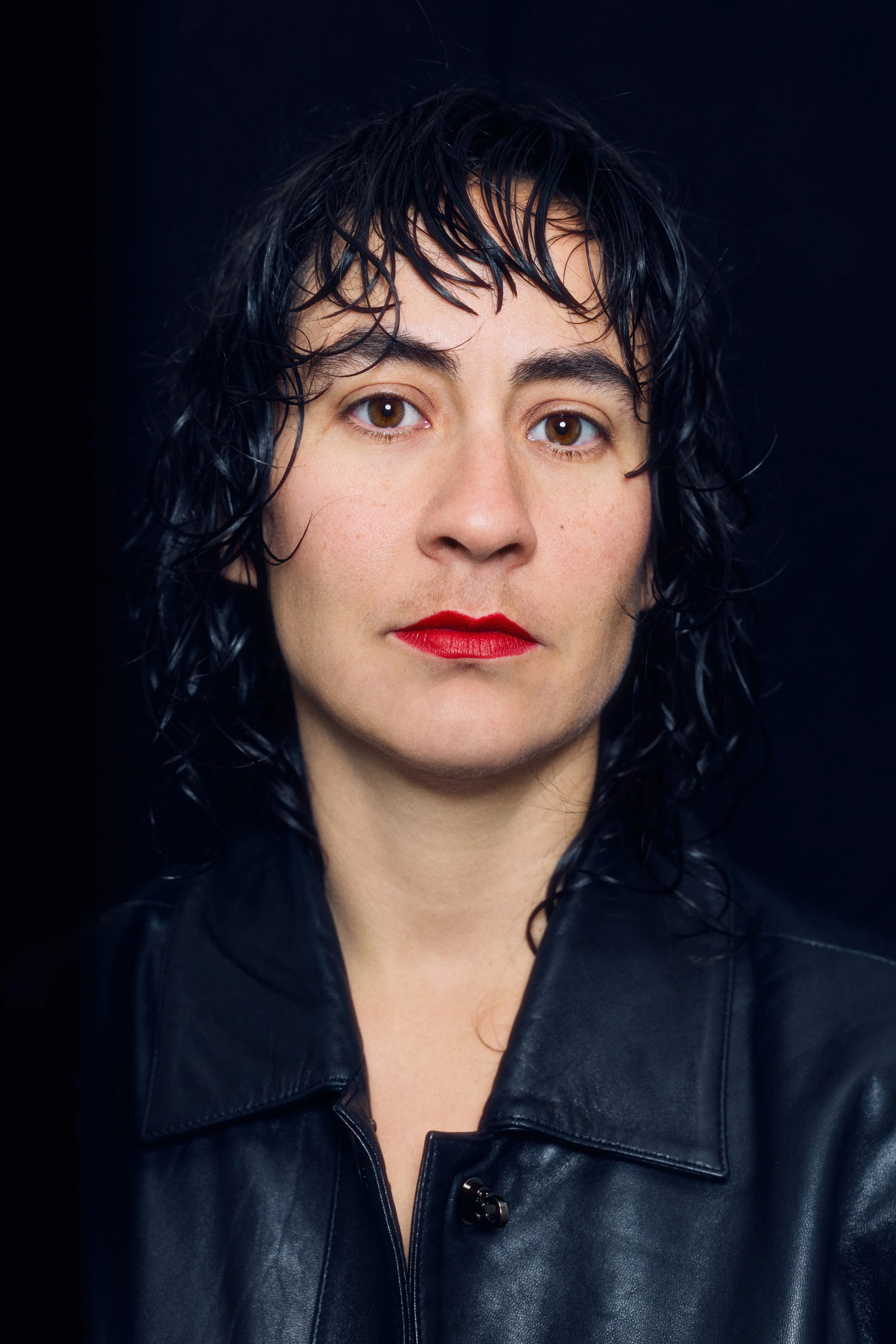Close-up of a woman with wet black hair, wearing a black leather jacket, red lipstick, and looking directly at the camera.