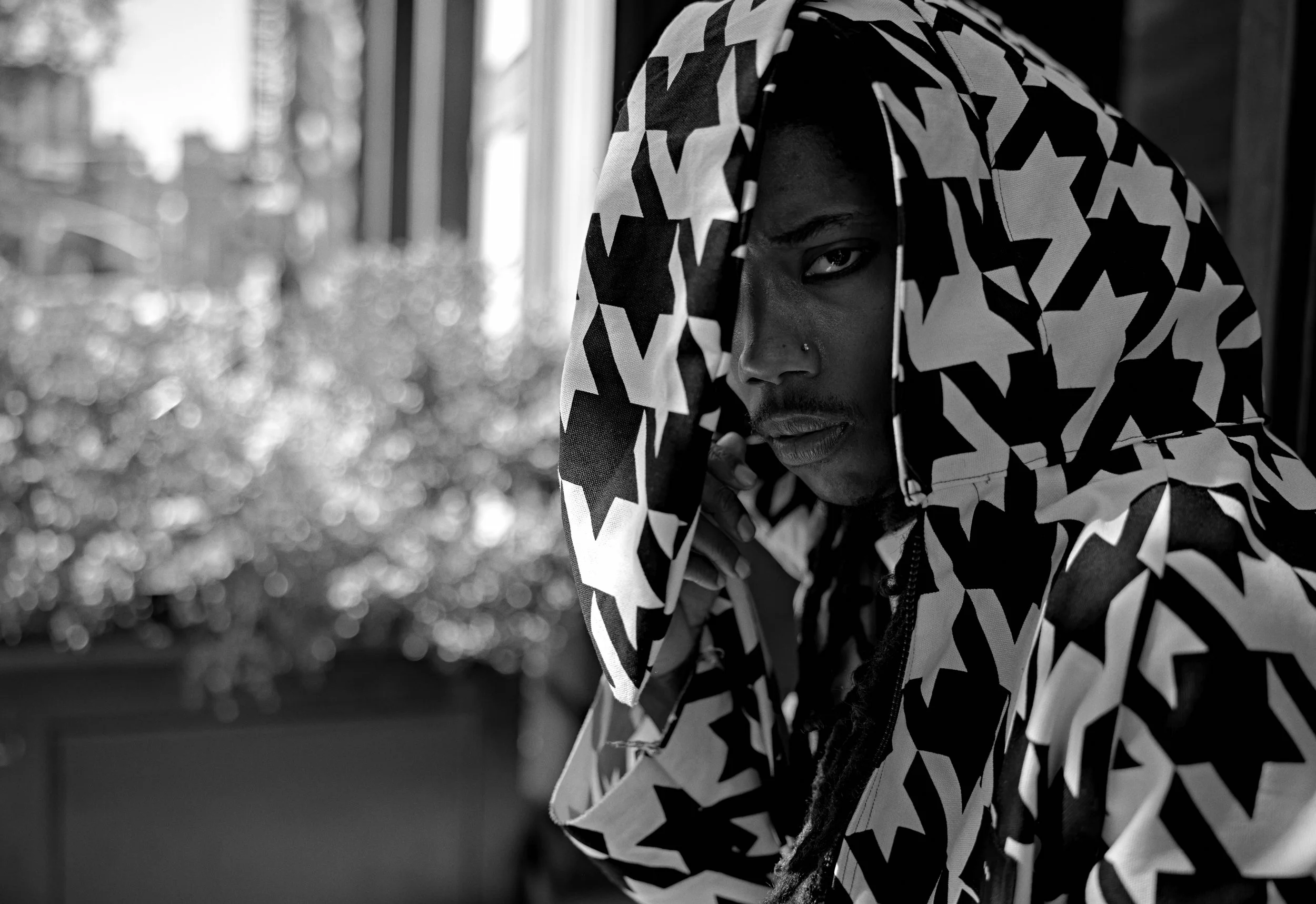 Black-and-white portrait of a person with long braids wearing a bold geometric-patterned hooded garment by a window