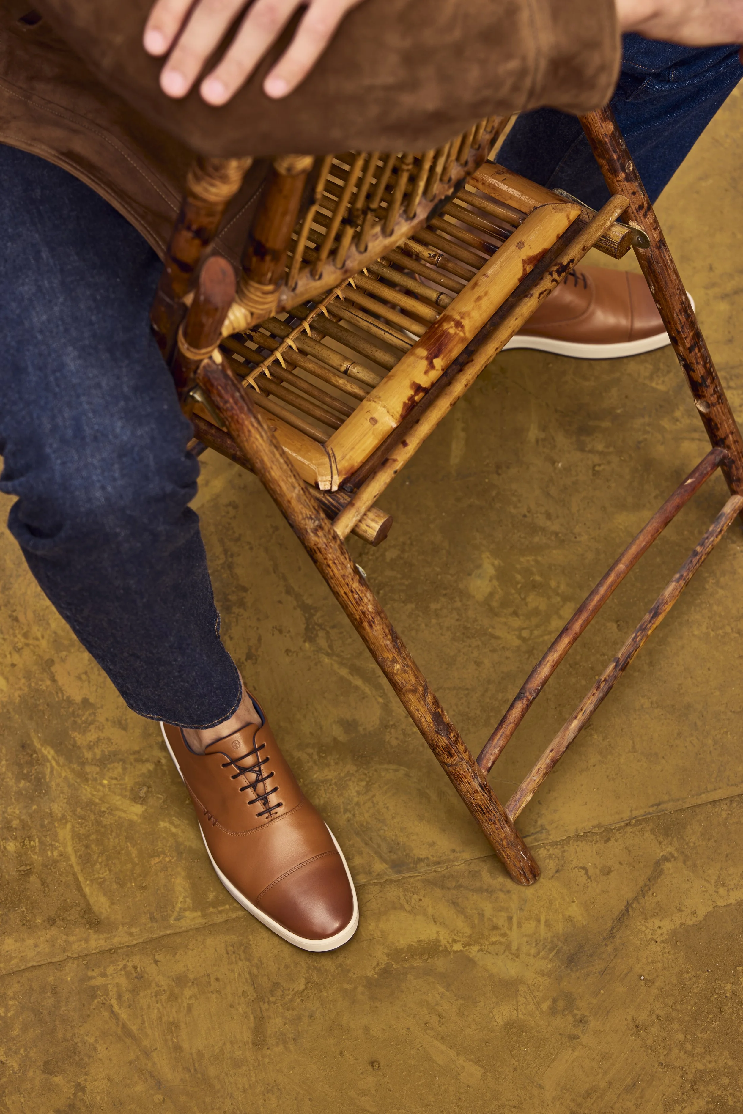 Overhead view of a person sitting on a wooden chair with a bamboo seat, wearing brown leather shoes, dark blue jeans, and a brown jacket, with one hand resting on their lap, on a yellowish-brown floor.