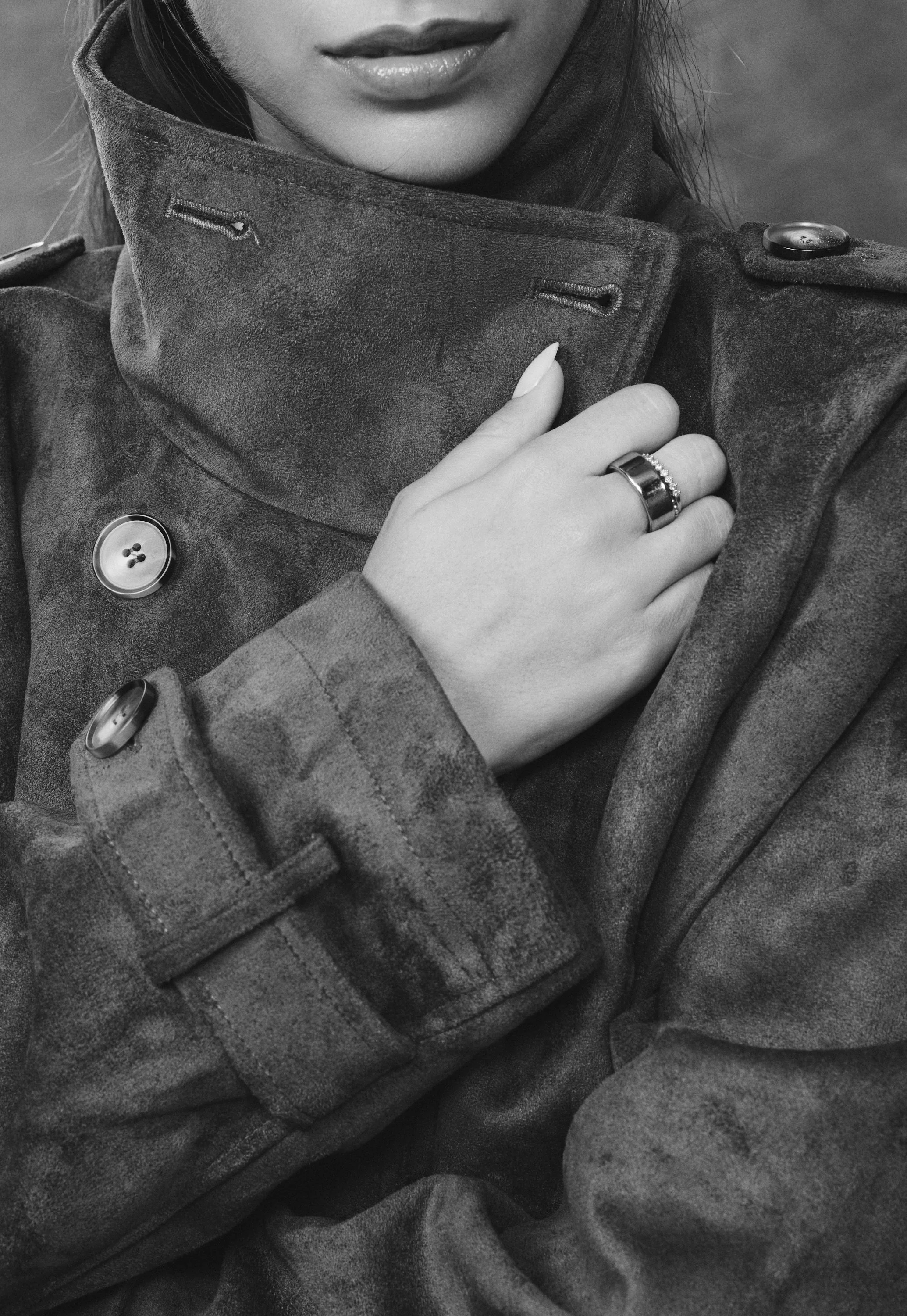 Black-and-white close-up of a person wearing a wool coat and scarf, with a hand adjusting the collar and displaying rings.