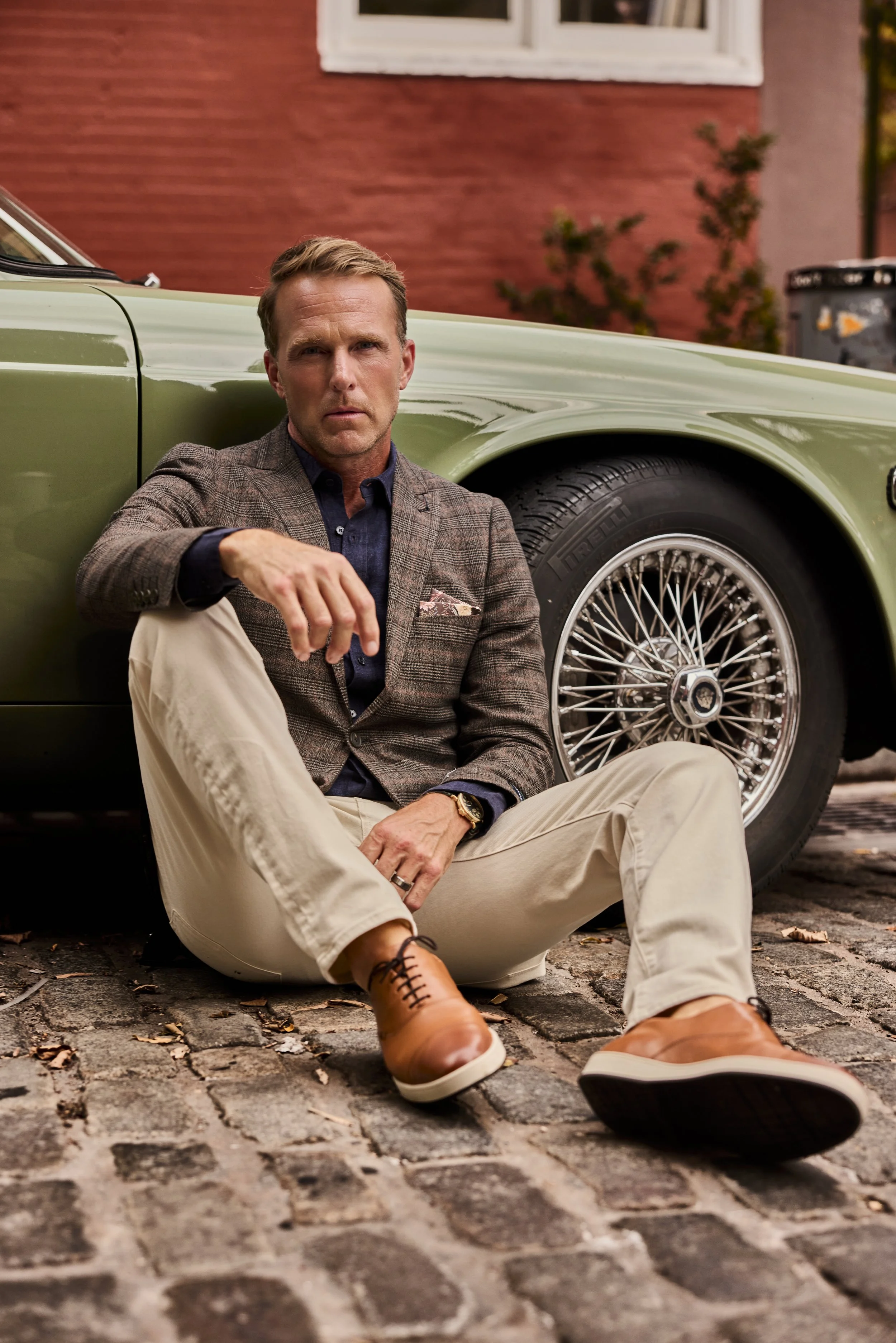 A man sitting on cobblestone street near a vintage green car, dressed in smart casual attire, with a brick building in the background.