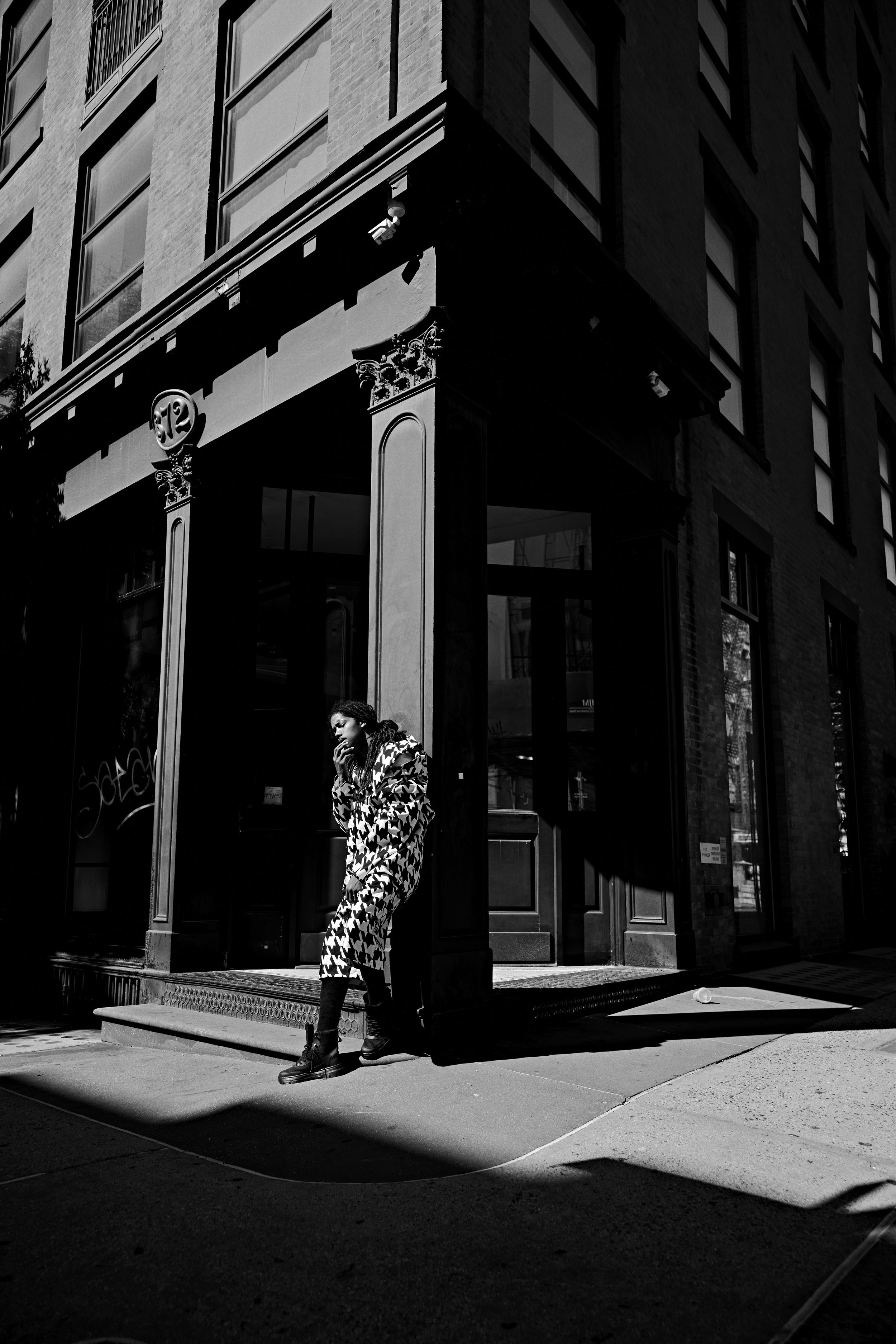 Black-and-white street photo of a woman in a bold patterned coat and boots walking past an ornate building entrance.