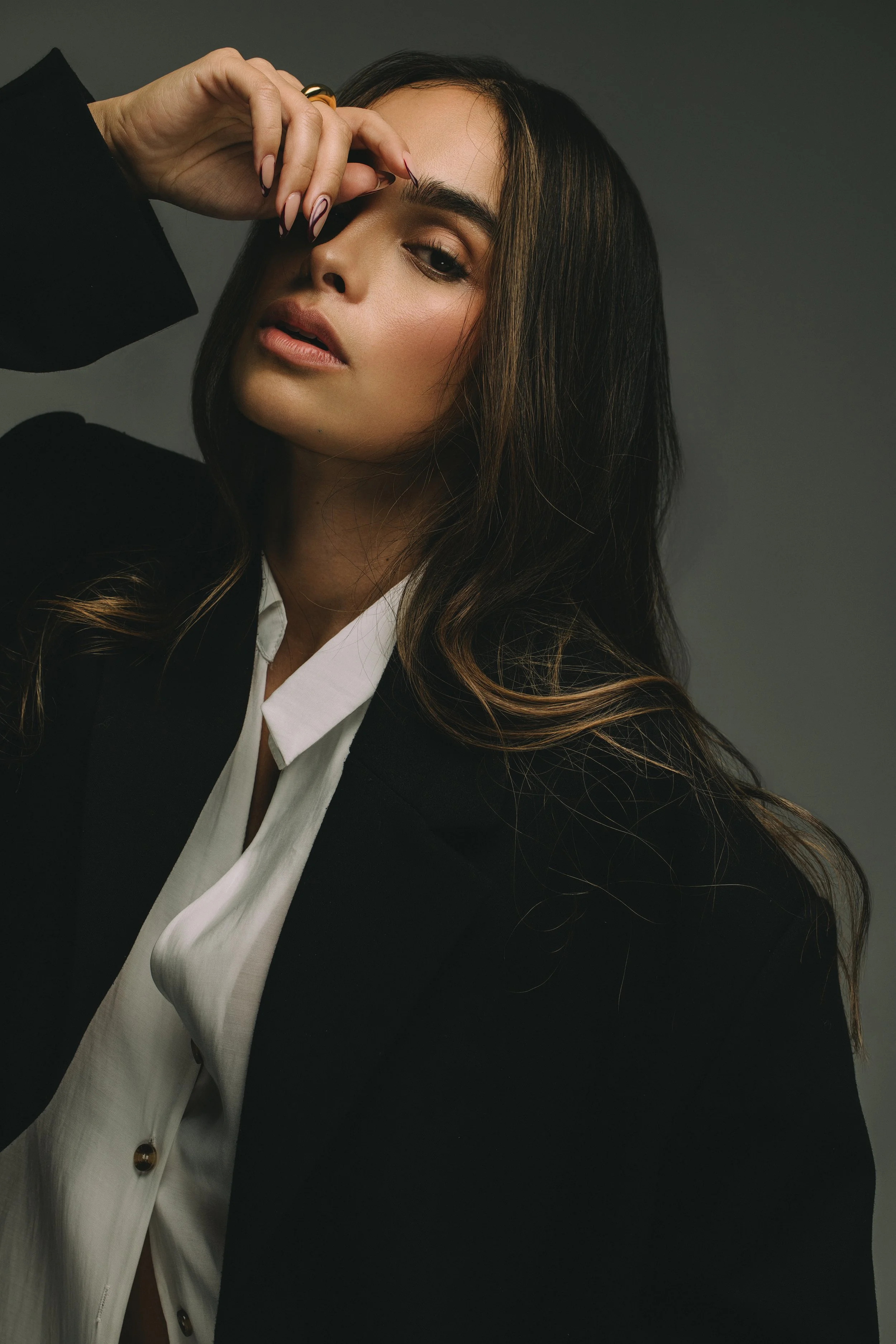 Close-up portrait of a woman with long dark hair, dramatic eye makeup, and manicured nails wearing a black blazer over a white shirt.