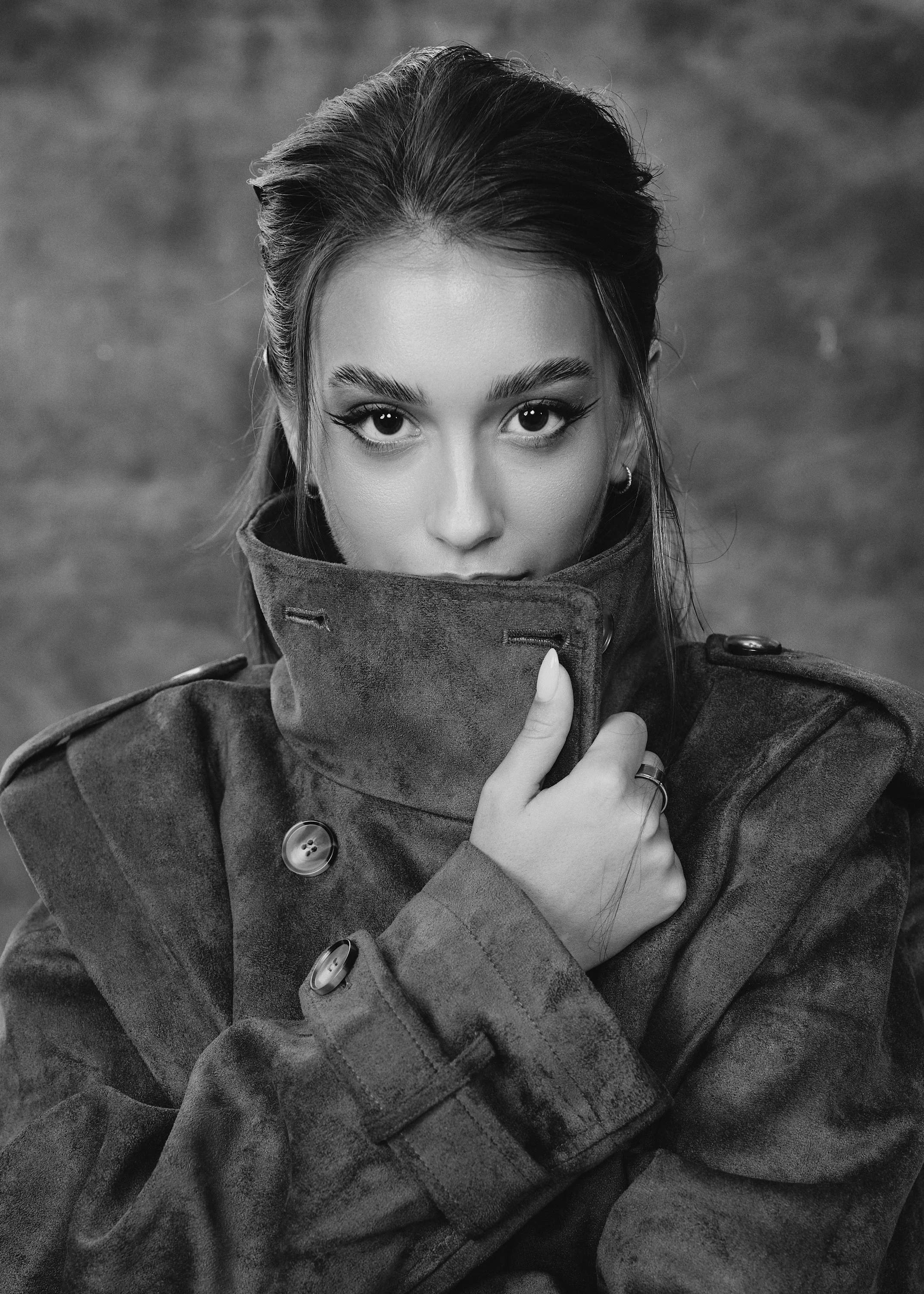 Black-and-white close-up portrait of a woman wearing a high-collared coat and holding the collar over her mouth