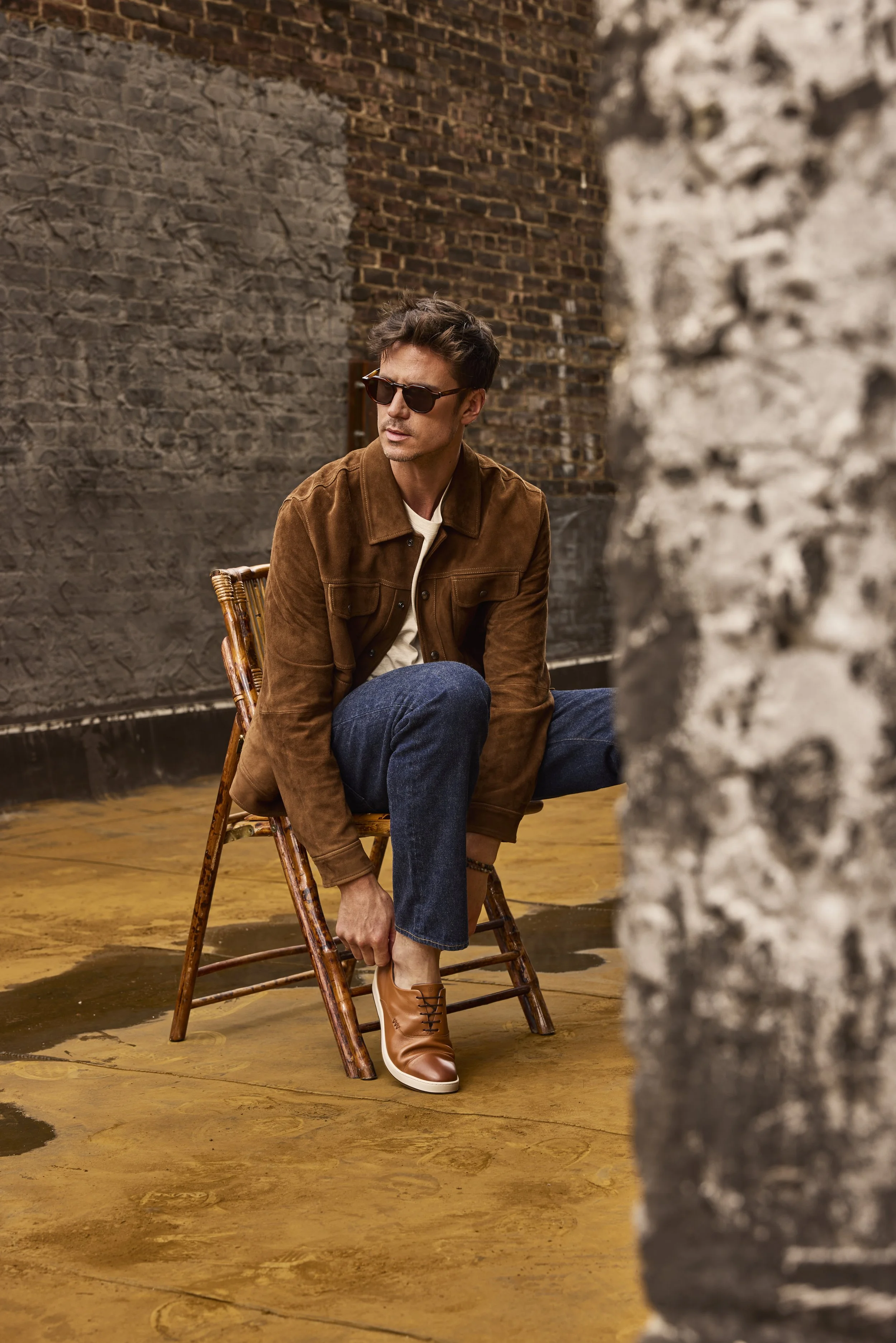 A man sitting on a rattan chair outdoors, adjusting his brown leather shoe. He's wearing sunglasses, a brown jacket, a white shirt, and blue jeans, with a brick wall and a rough gray wall in the background.