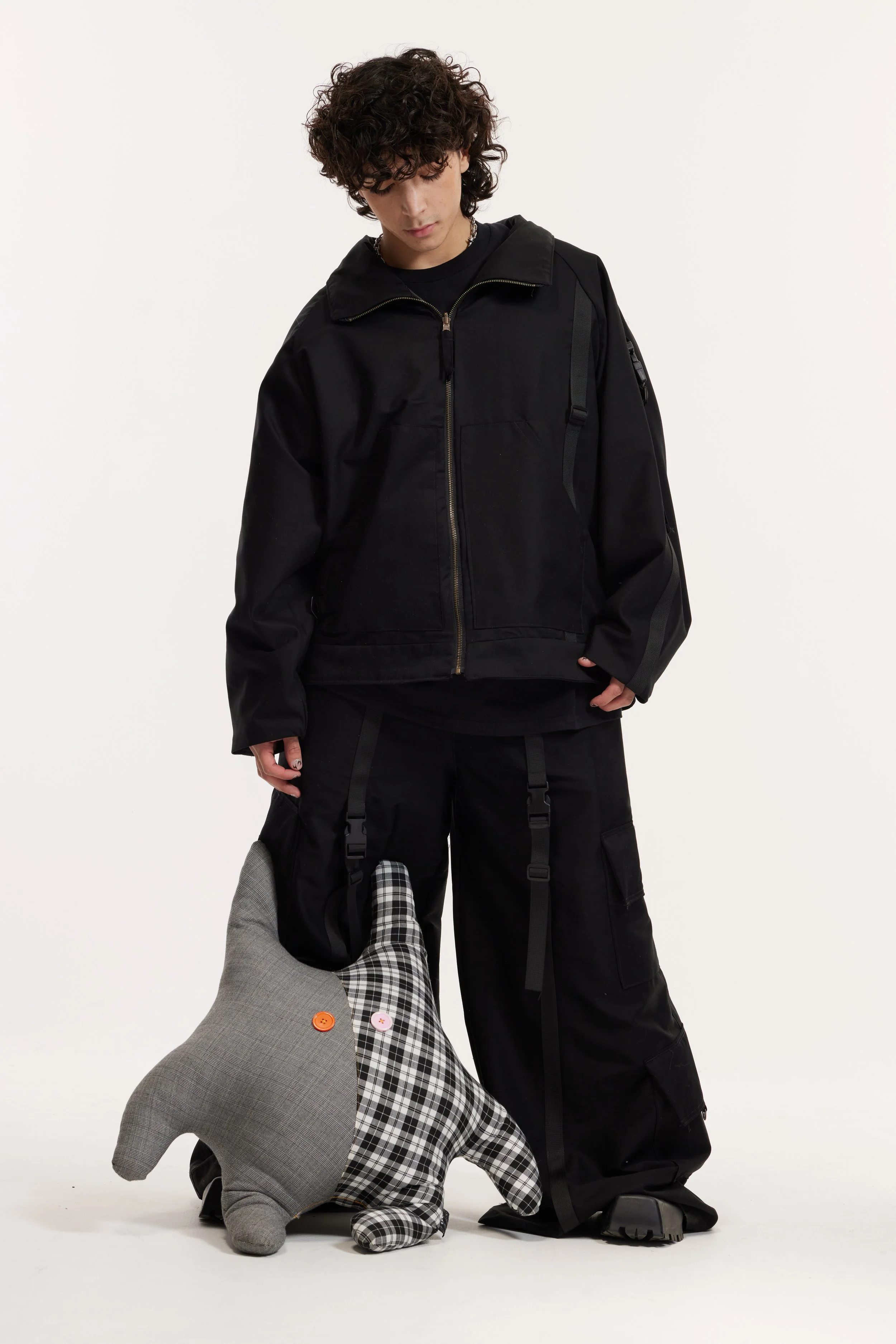 Model wearing a black utility jacket and cargo pants, standing beside two plaid plush dogs.