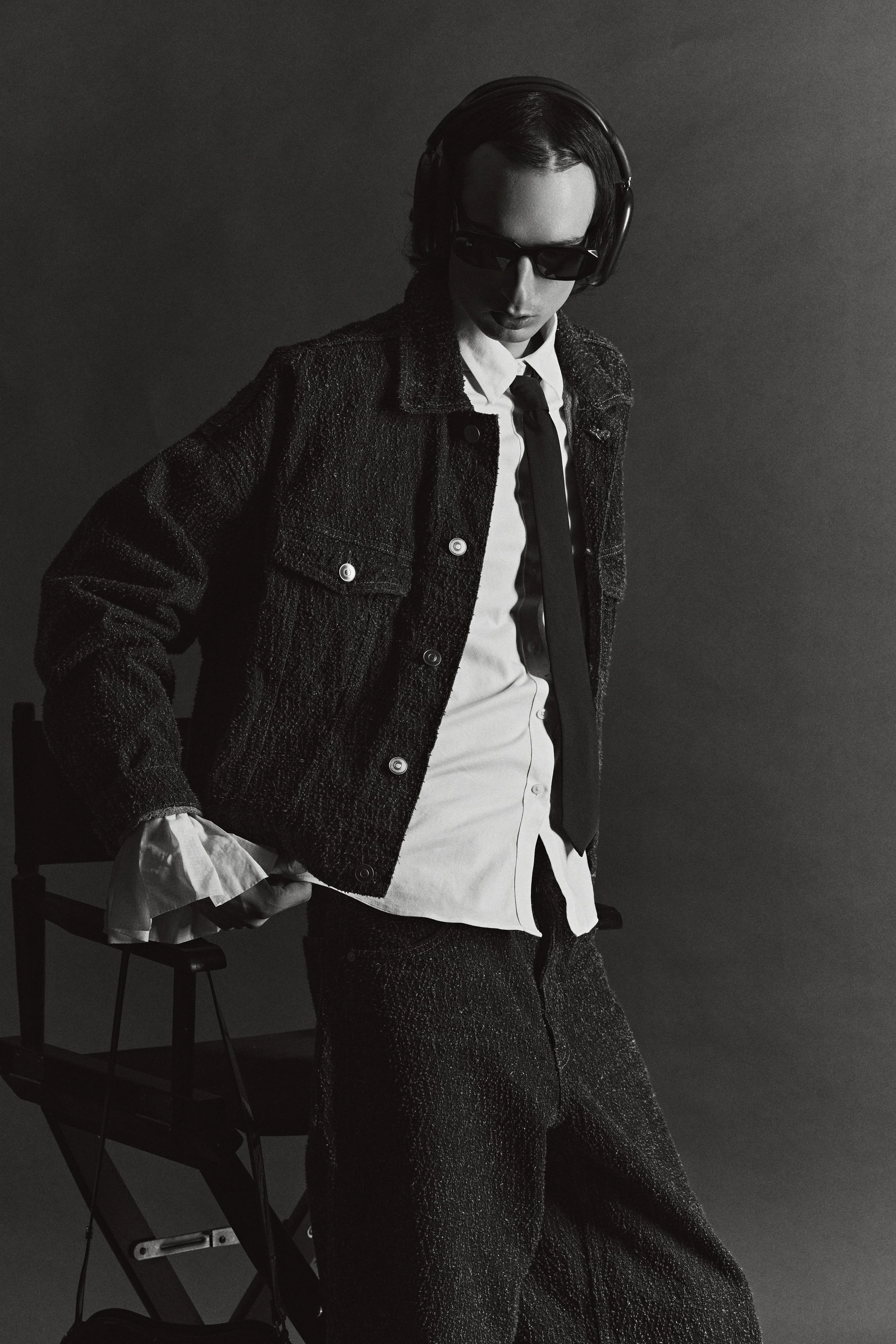 Black-and-white portrait of a person wearing sunglasses, over-ear headphones, a textured cardigan, white shirt, and tie against a dark background