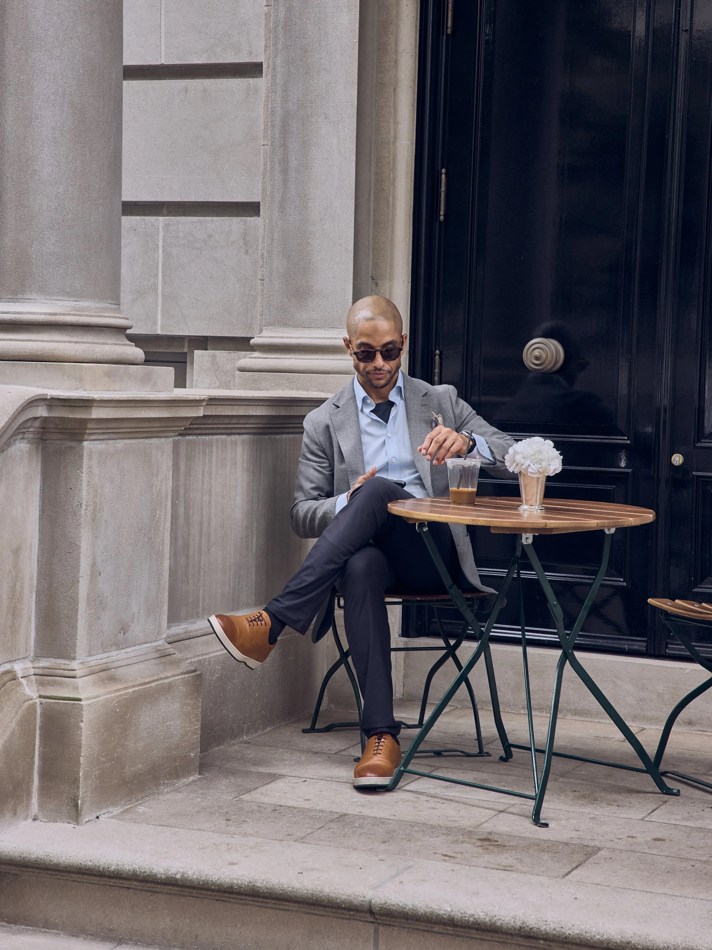 Man in sunglasses sitting at outdoor cafe table with drink and flowers outside stone building