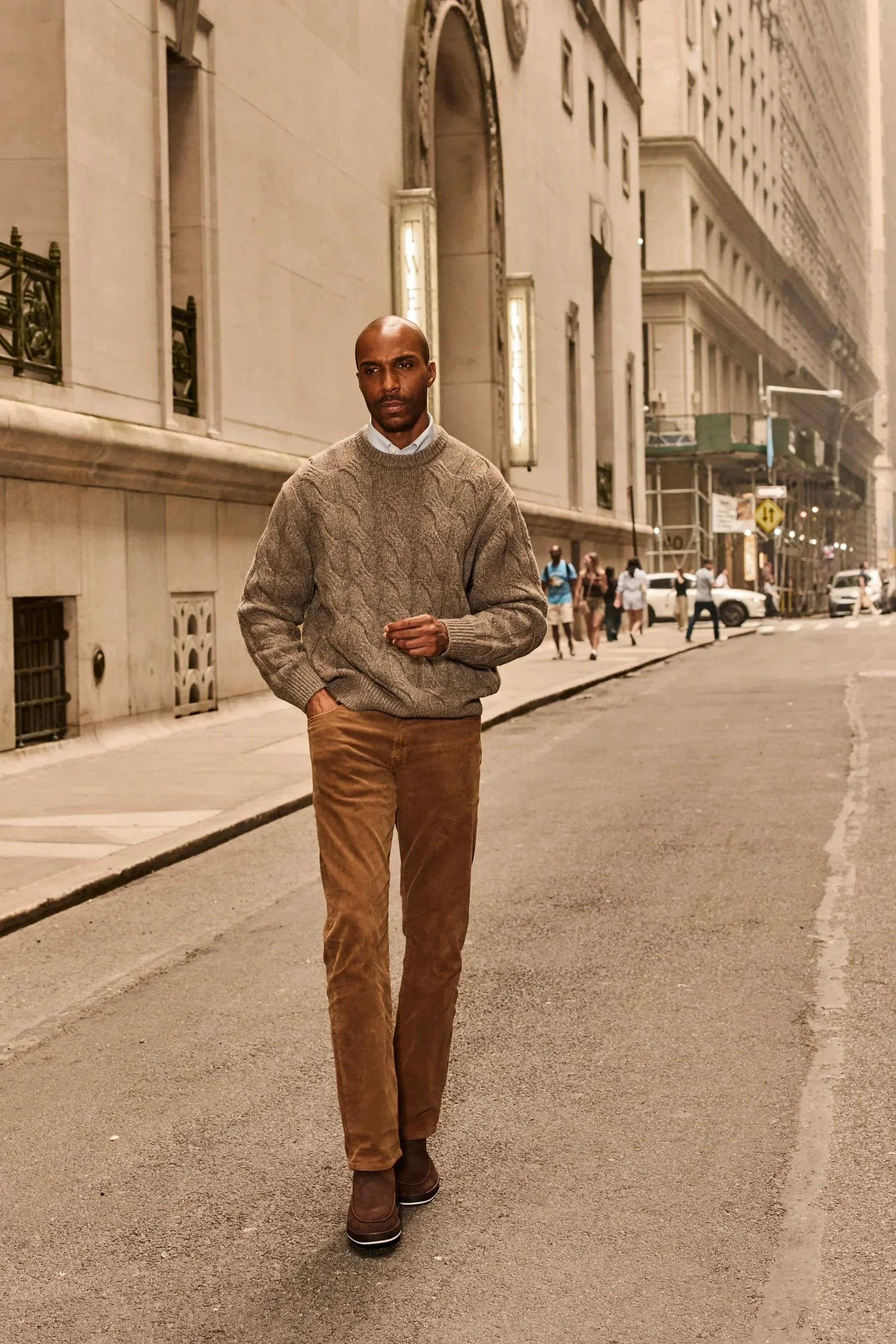 Bald man in a cable-knit sweater and brown pants walking through a downtown city street