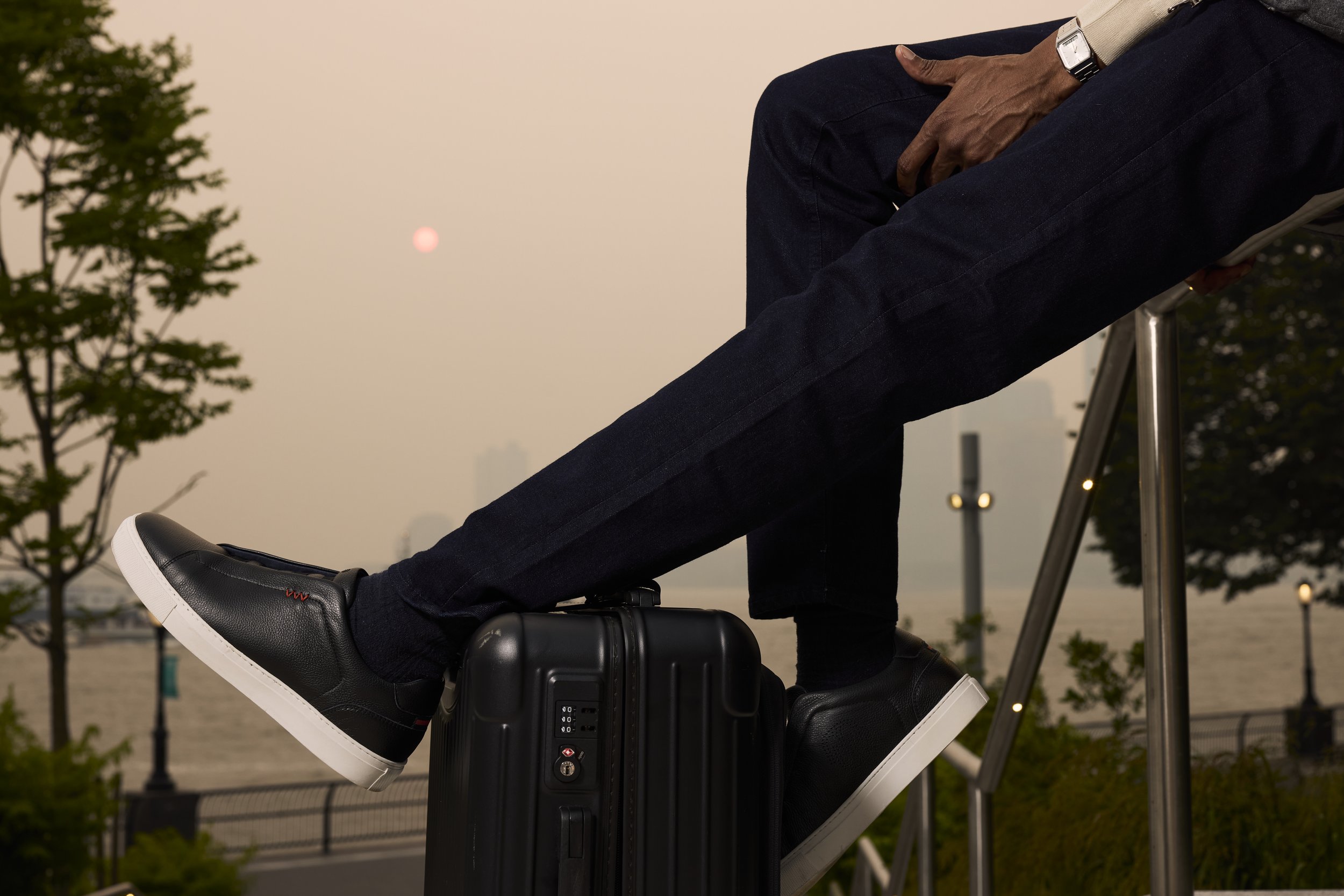 Traveler in dark jeans and black sneakers resting feet on a black suitcase outdoors at sunset