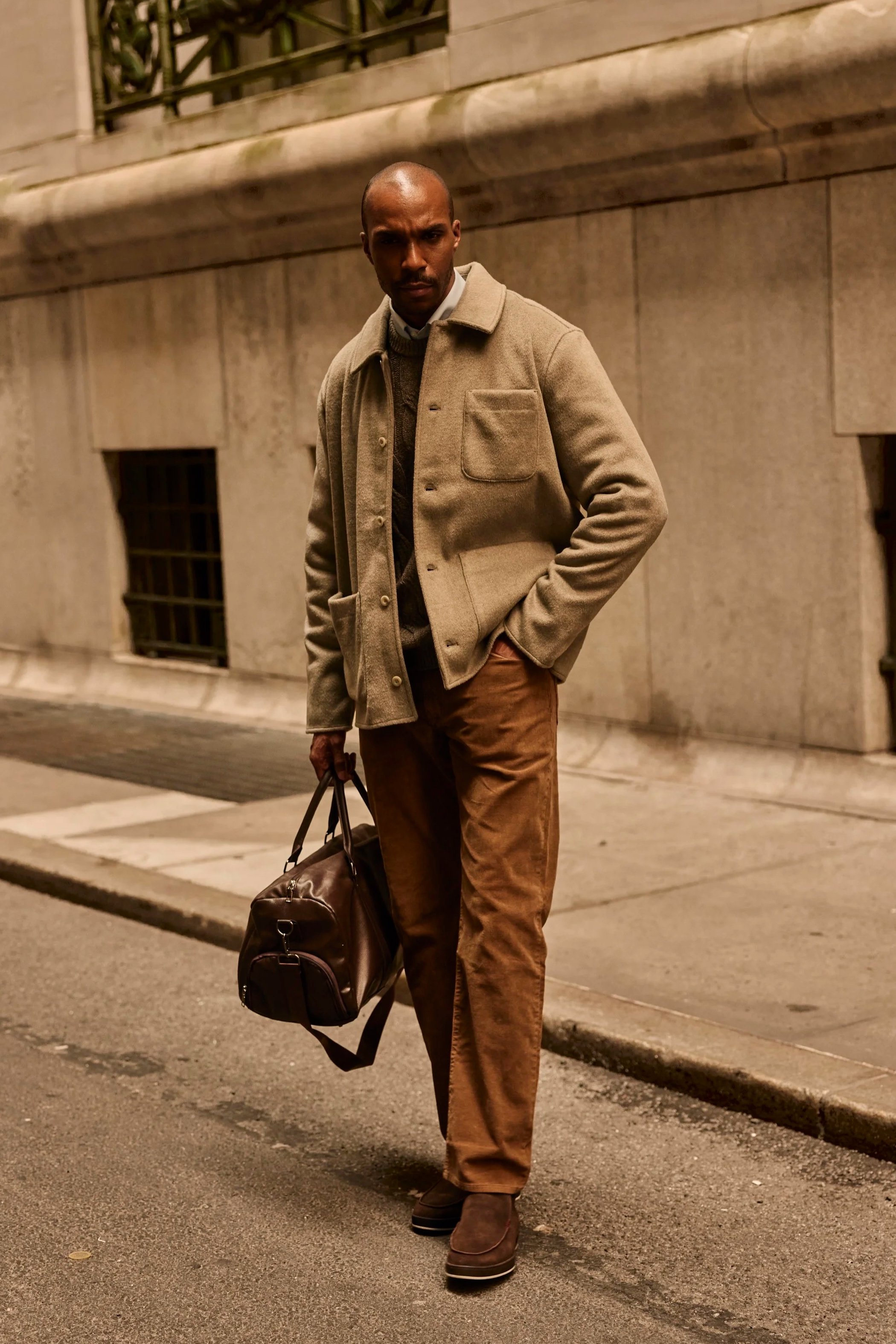 Bald man in a tan wool jacket and brown pants carrying a leather duffel bag on a city street