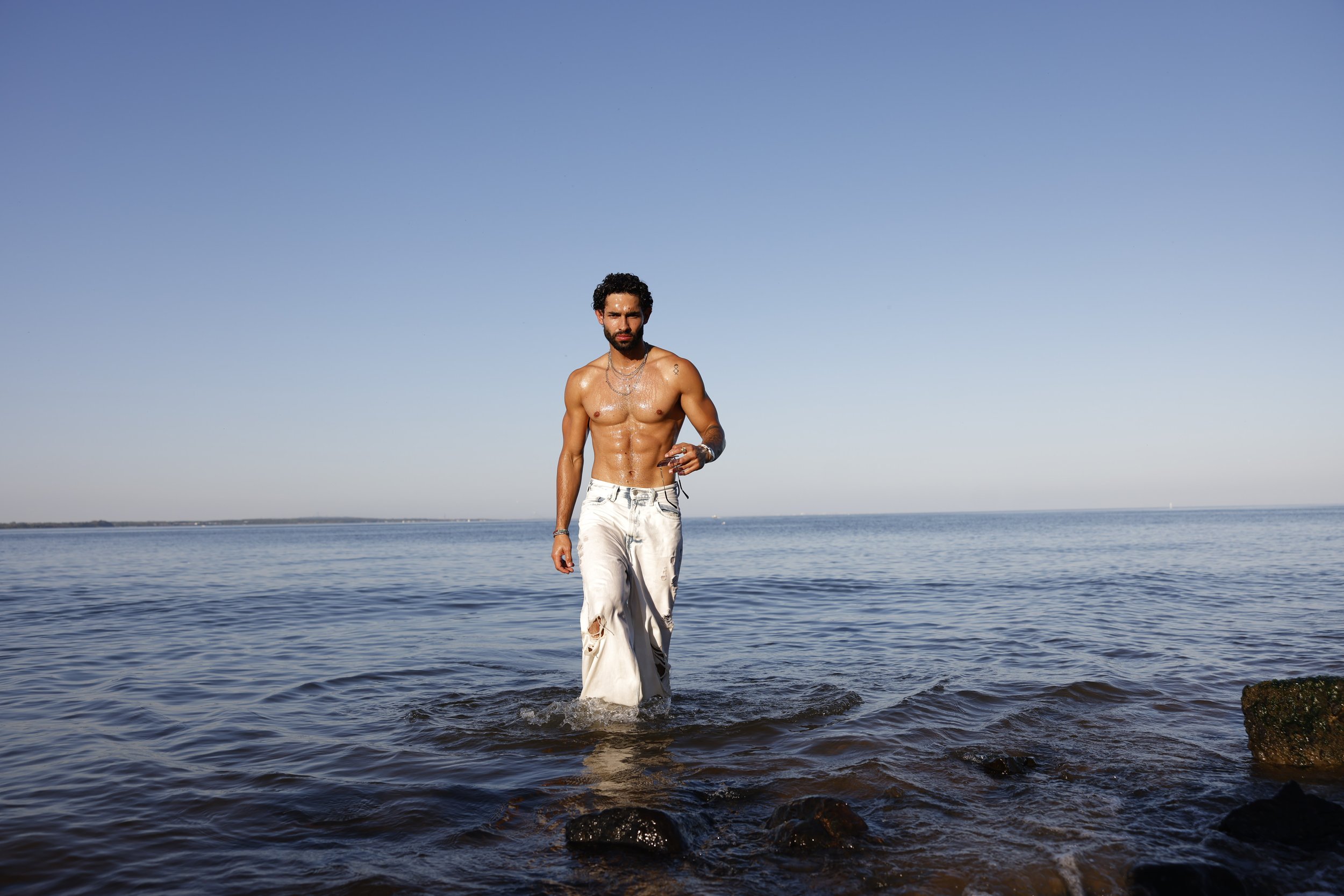 Shirtless man wearing white pants walks through shallow ocean water under a clear blue sky