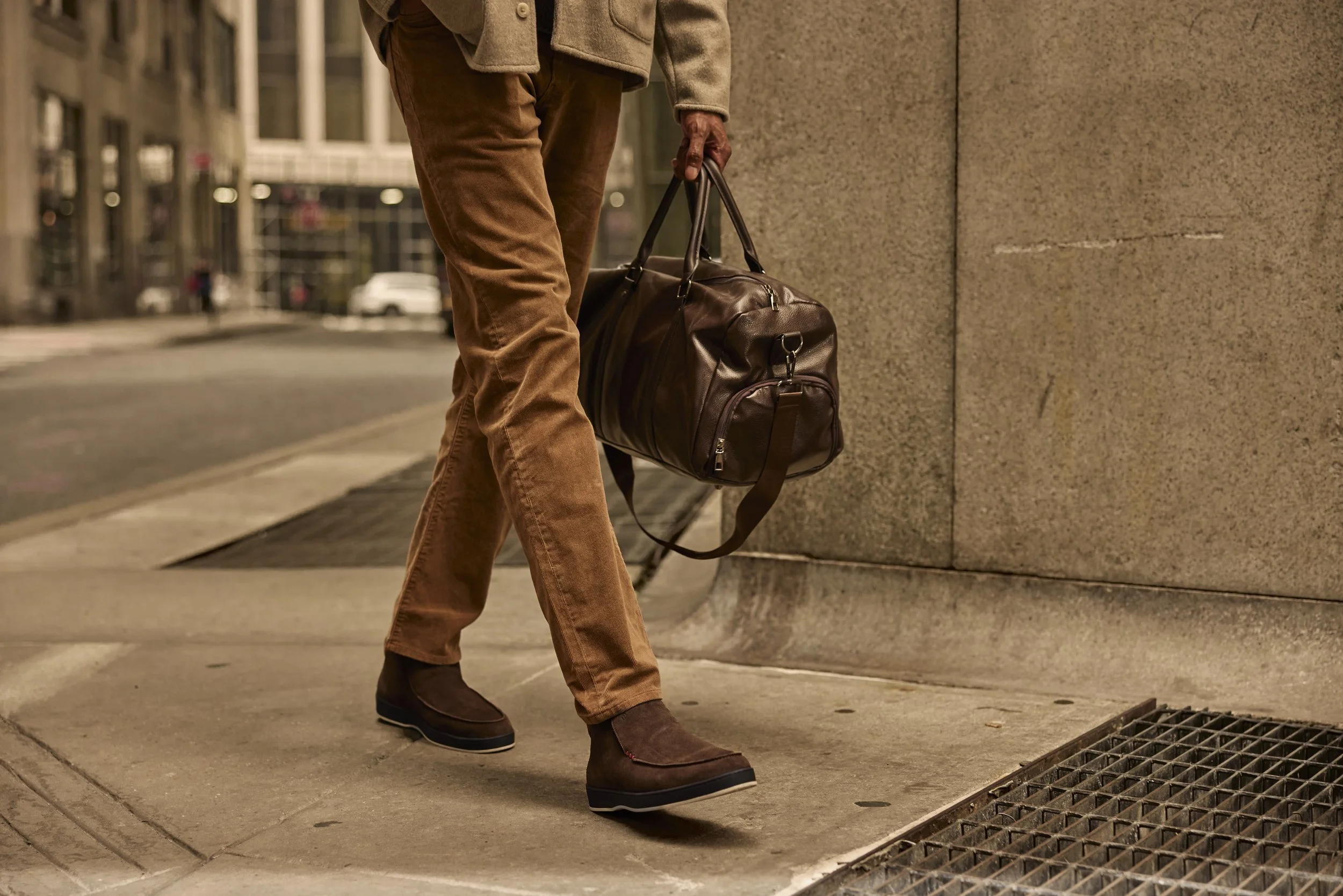 Person walking on a city sidewalk carrying a brown leather duffel bag