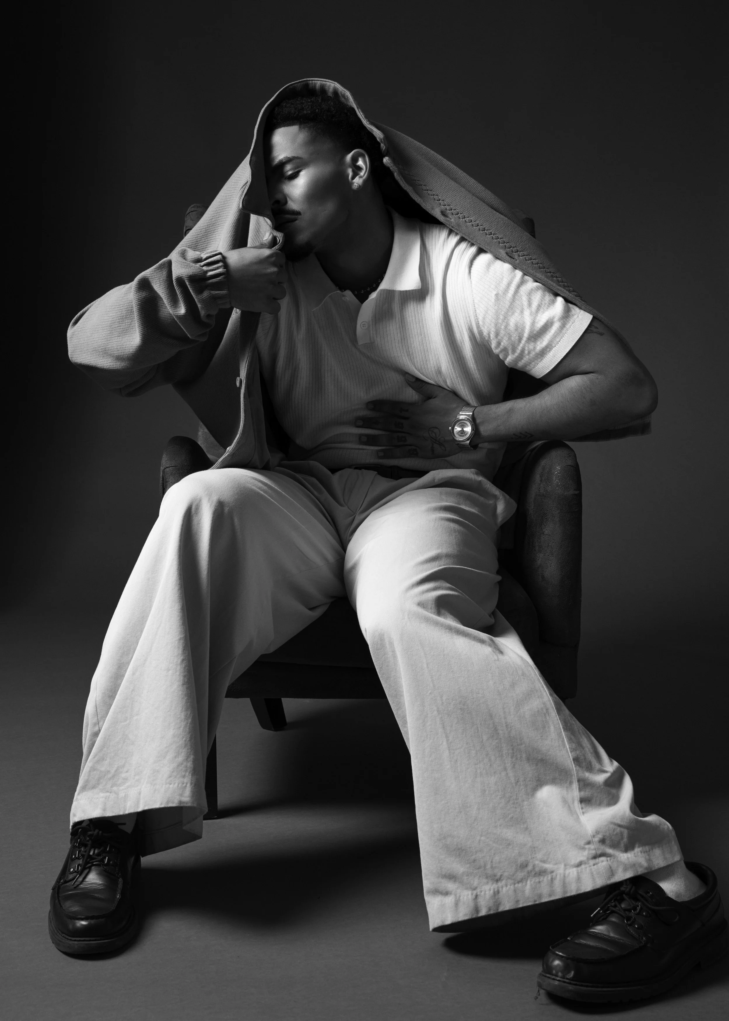 Black-and-white portrait of a man sitting in a chair wearing a hooded jacket, white shirt, wide-leg pants, and black shoes.