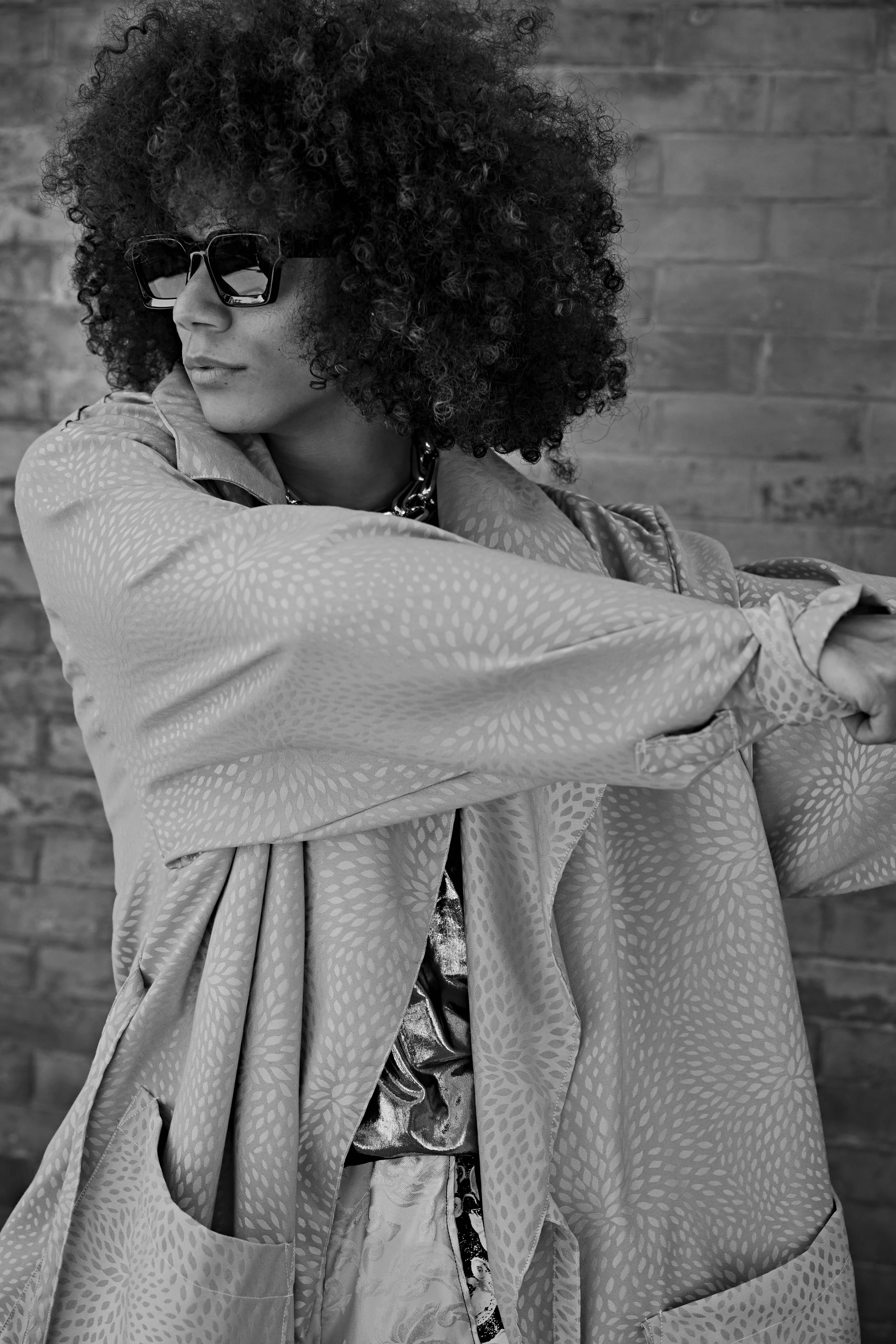 Black-and-white portrait of a person with curly hair and sunglasses wearing a patterned blazer against a brick wall.