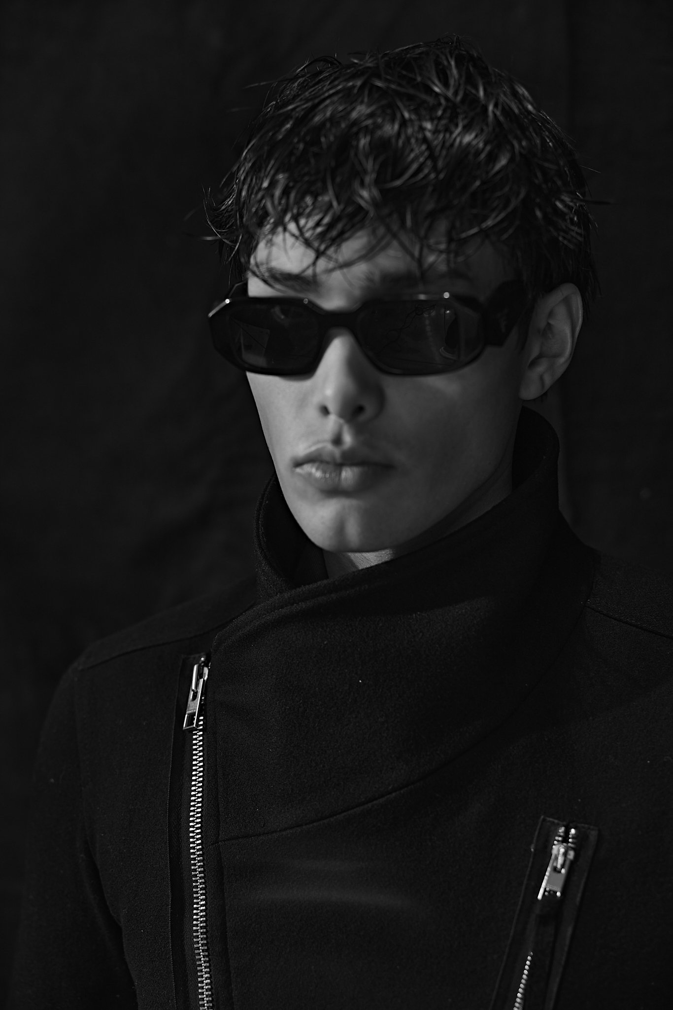 Black-and-white portrait of a person with tousled hair, dark sunglasses, and a black zip-up jacket against a dark background