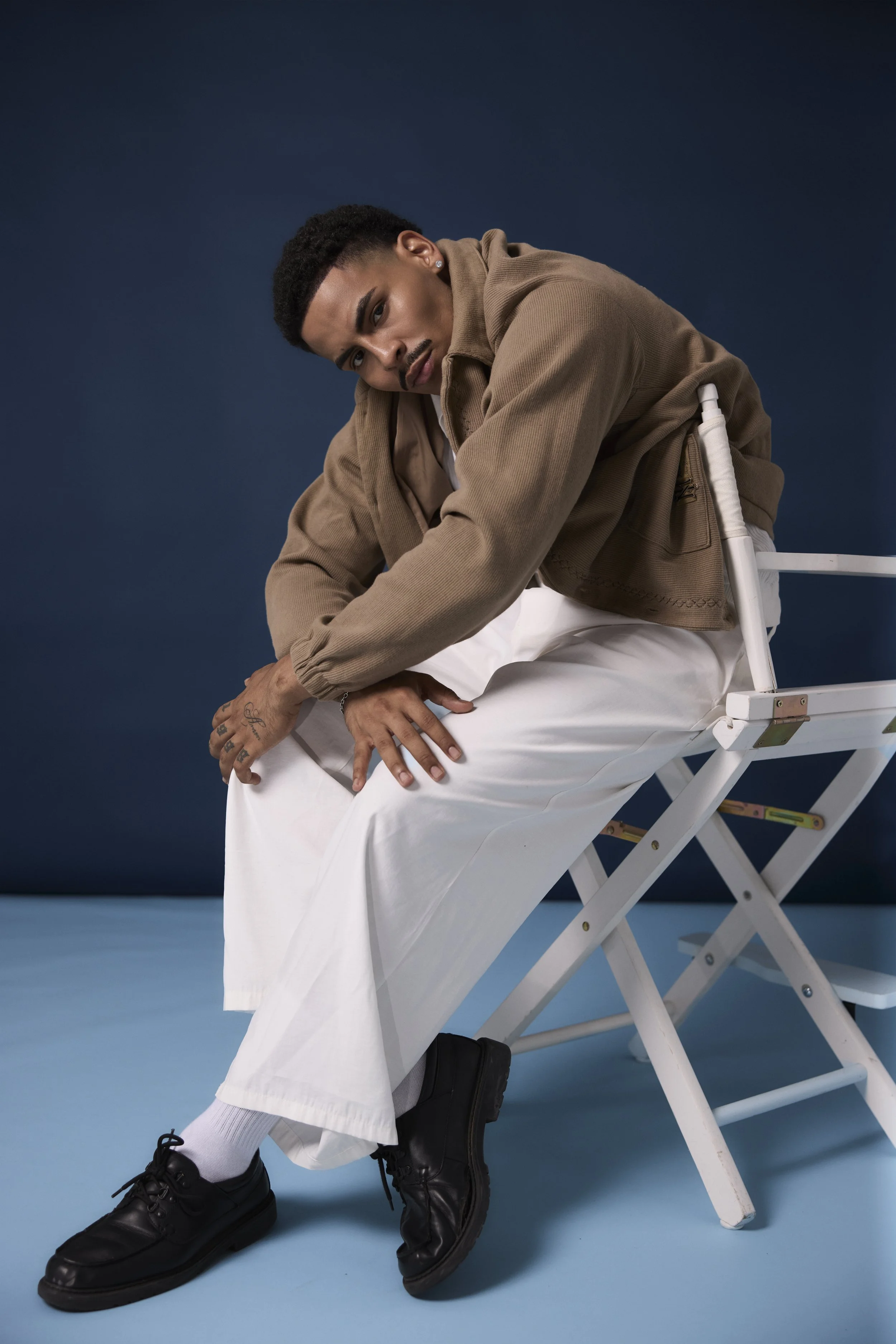 Young man in a tan jacket and white pants sitting backward on a white folding chair against a dark blue background