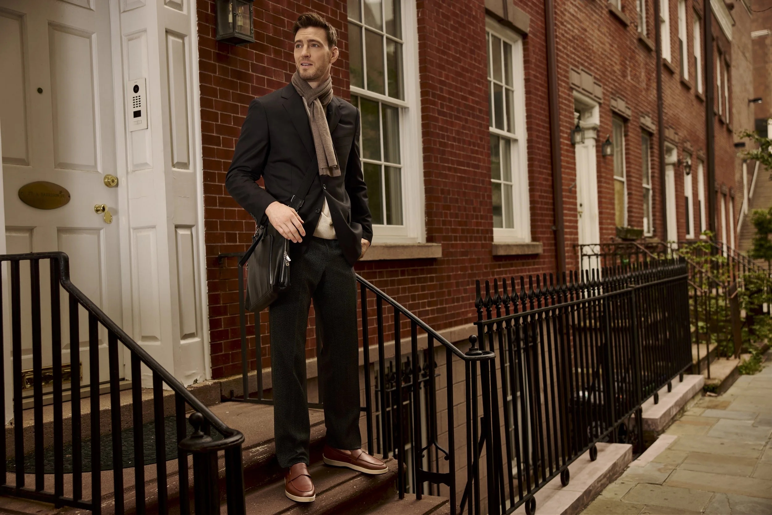Well-dressed man in a dark suit and scarf standing on brick townhouse steps with a leather briefcase.