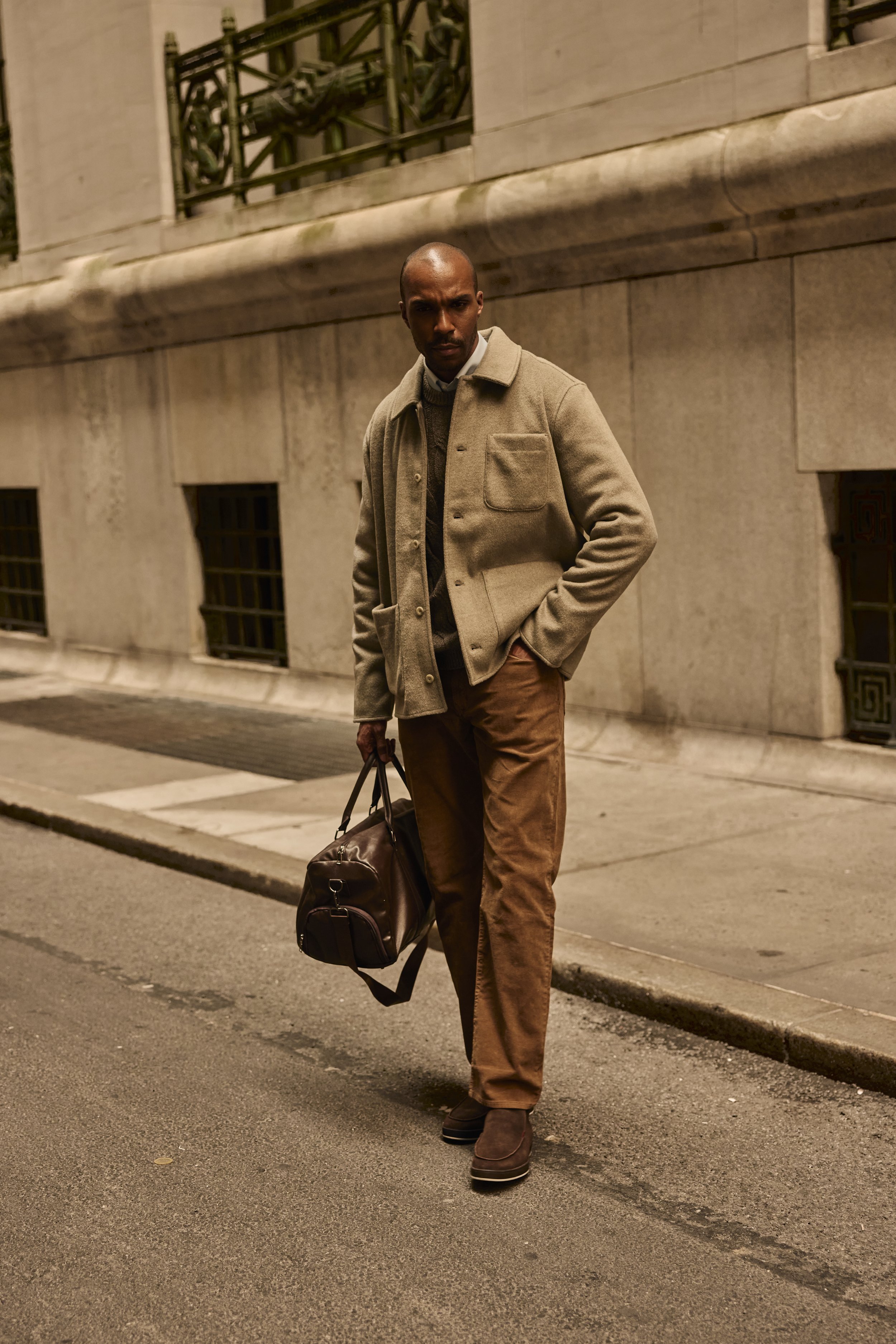 Bald man in a tan coat carrying a leather duffel bag on a city street