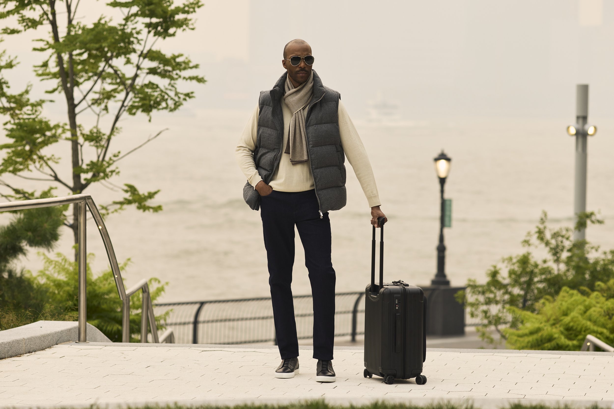 Man wearing sunglasses, scarf, and gray vest standing outdoors with a black rolling suitcase.