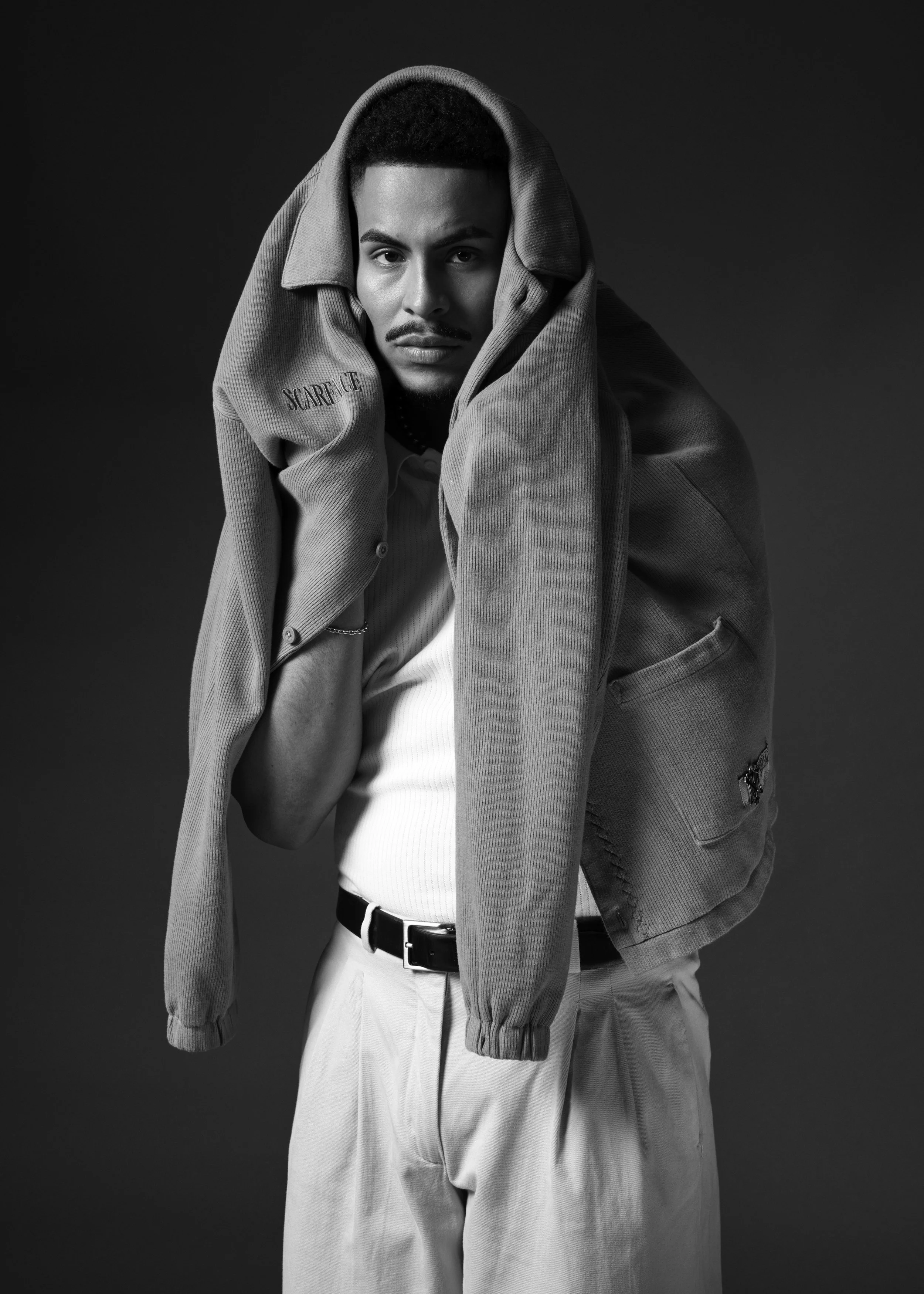 Black-and-white portrait of a man wearing a hooded cardigan, white shirt, chain necklace, and light trousers against a dark background.