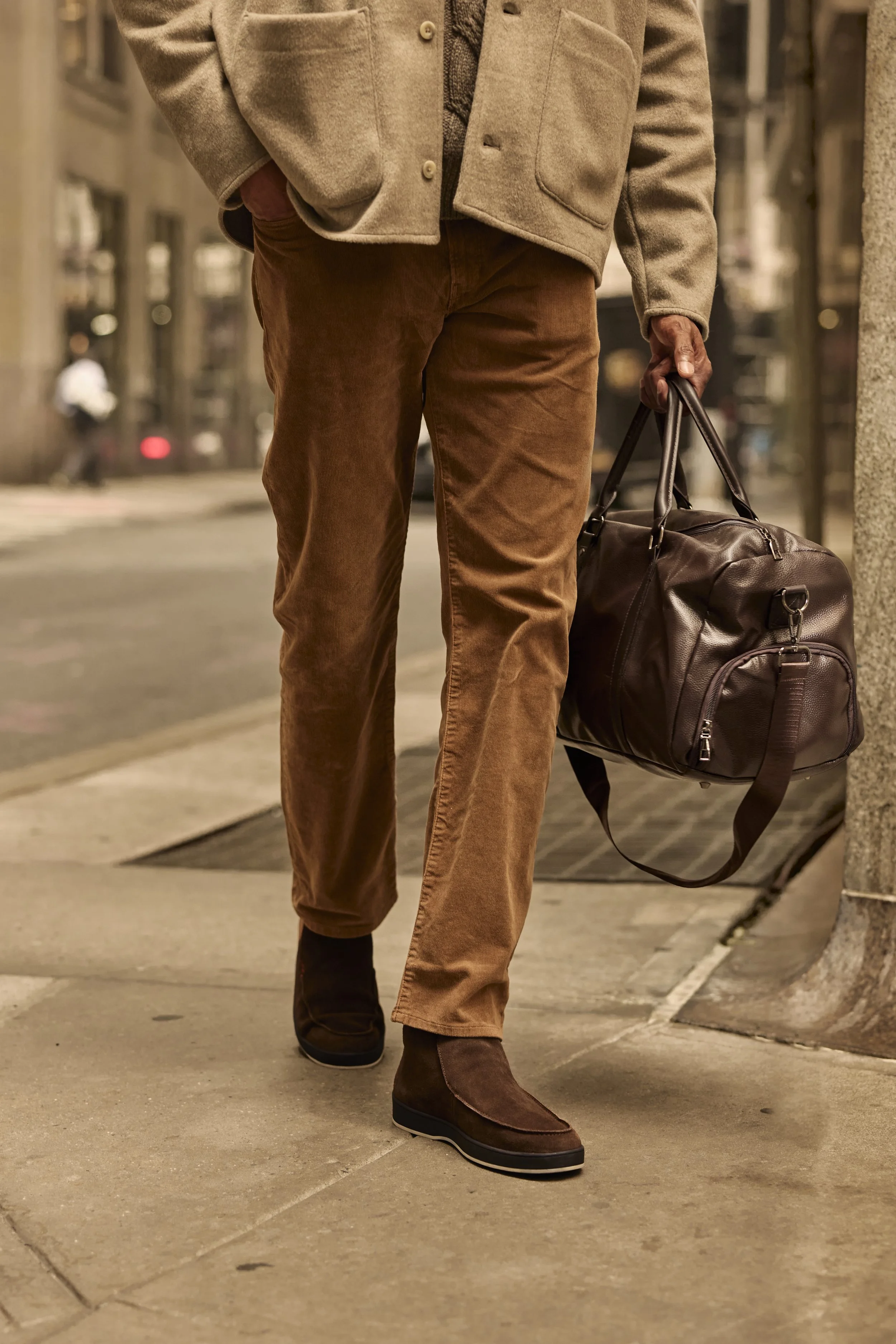 Person in a beige coat and brown corduroy pants walking with a brown leather duffel bag