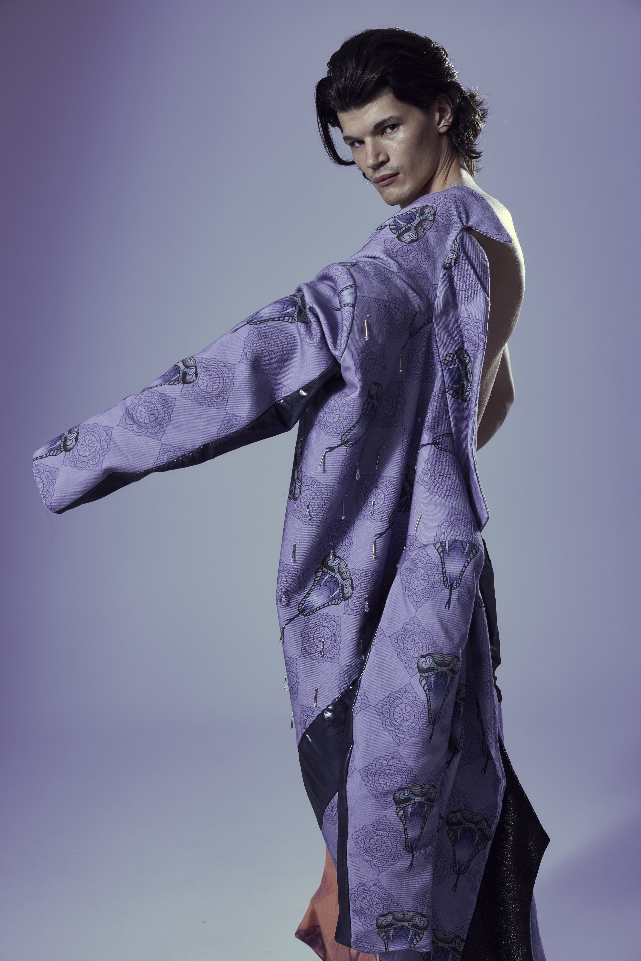 Male fashion model wearing a sheer purple patterned jacket with dangling embellishments against a lavender studio background