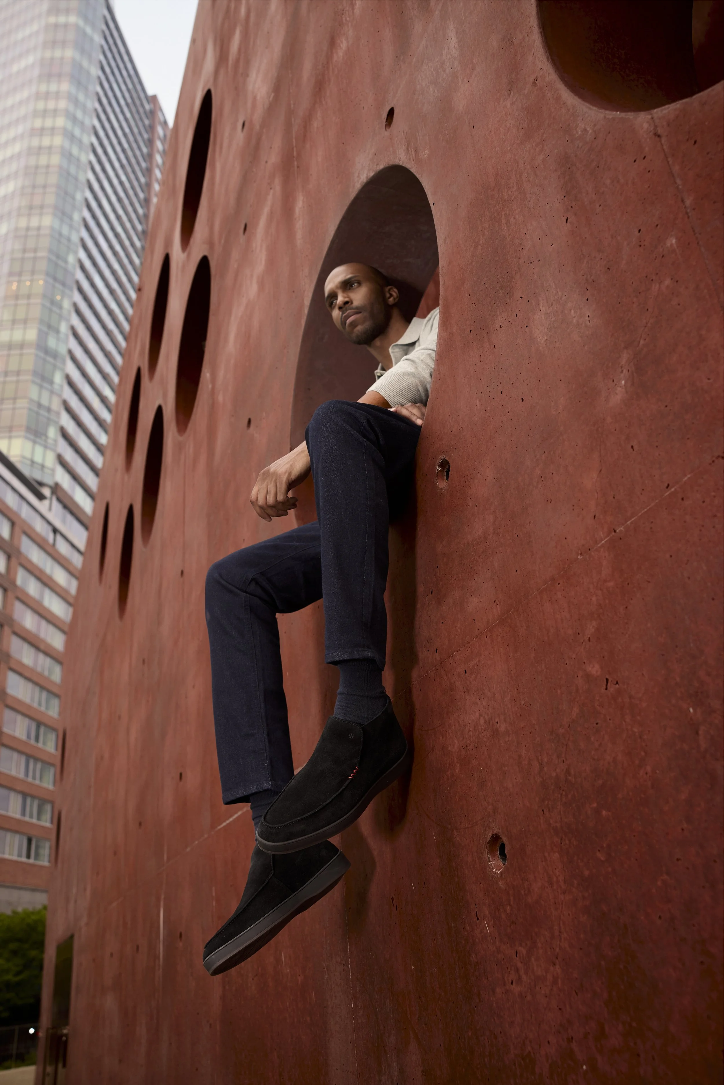 Man sitting inside a large rust-colored concrete sculpture with city buildings in the background