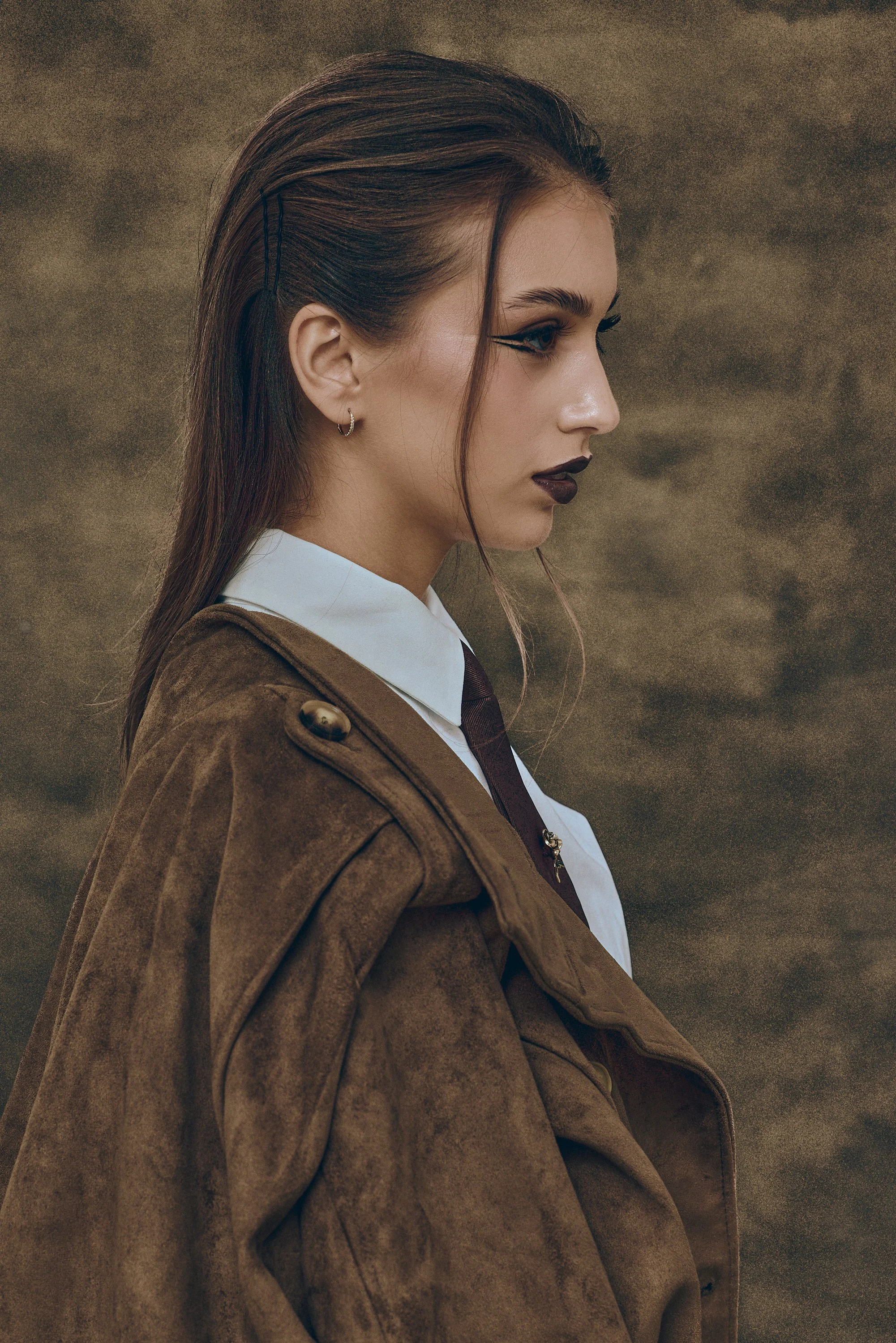 Side-profile portrait of a woman with dark hair, dramatic eyeliner, and dark lipstick wearing a brown coat and white collared shirt.