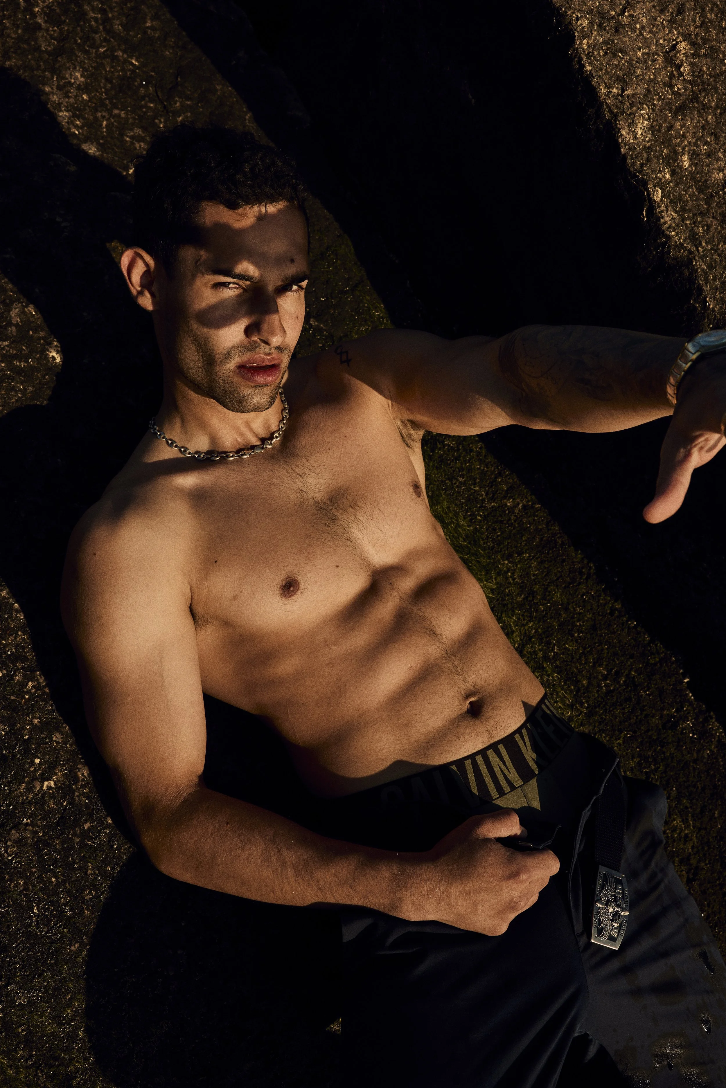 Shirtless muscular man reclining on a rocky surface, wearing a chain necklace and black pants in dramatic sunlight.