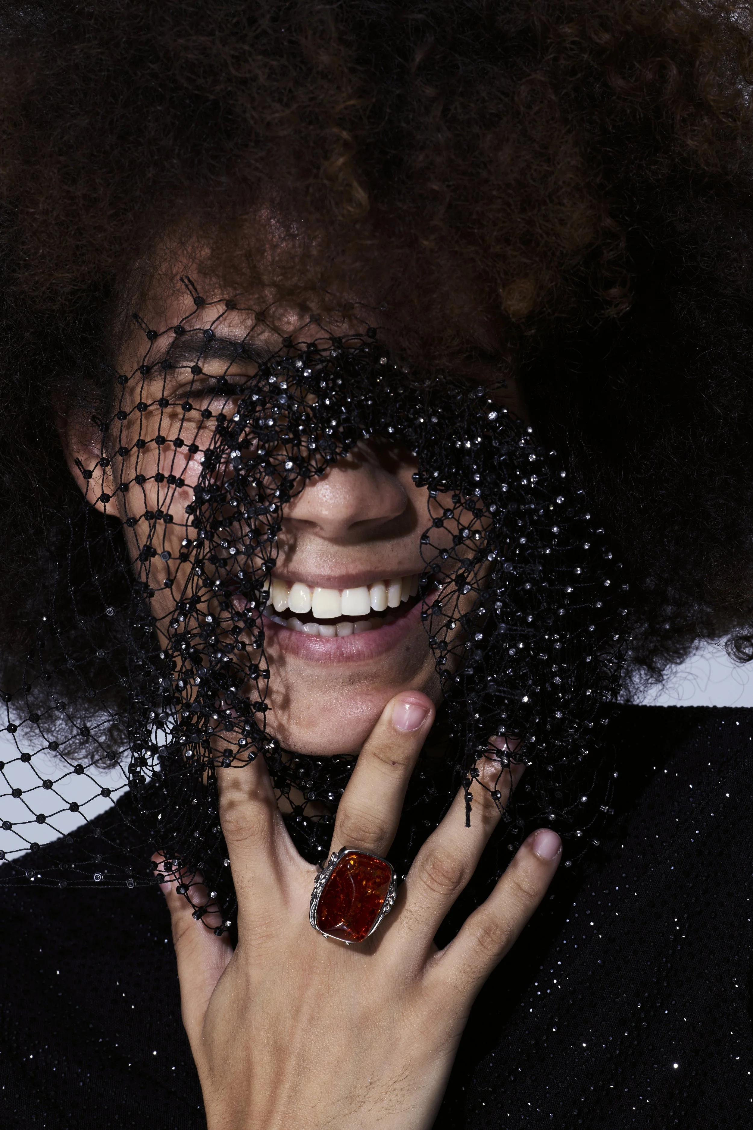 Close-up of a smiling person in black sheer beaded fabric, wearing a large amber gemstone ring.