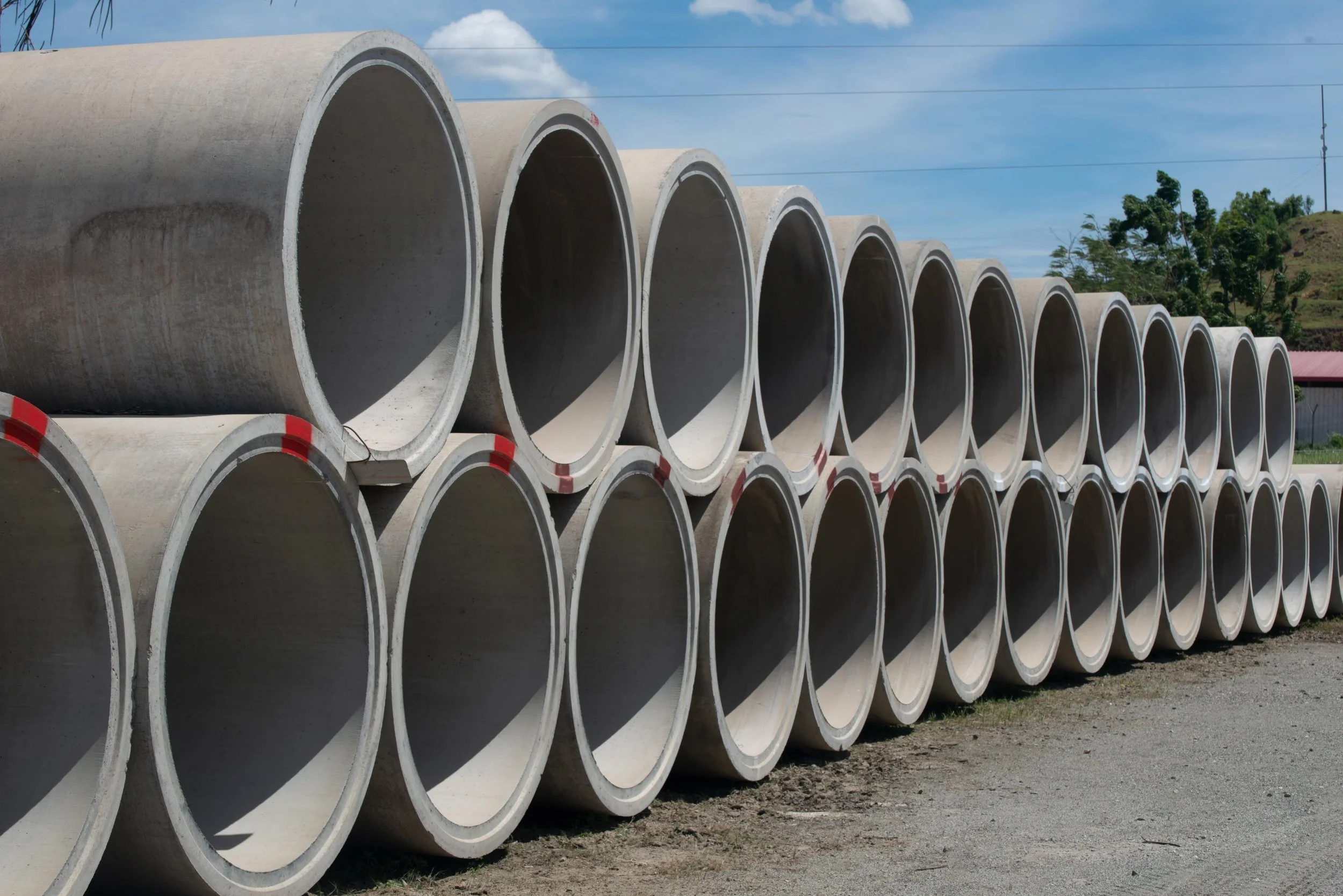 Multiple large concrete pipes stacked outdoors on a gravel surface, with trees and a blue sky with clouds in the background.