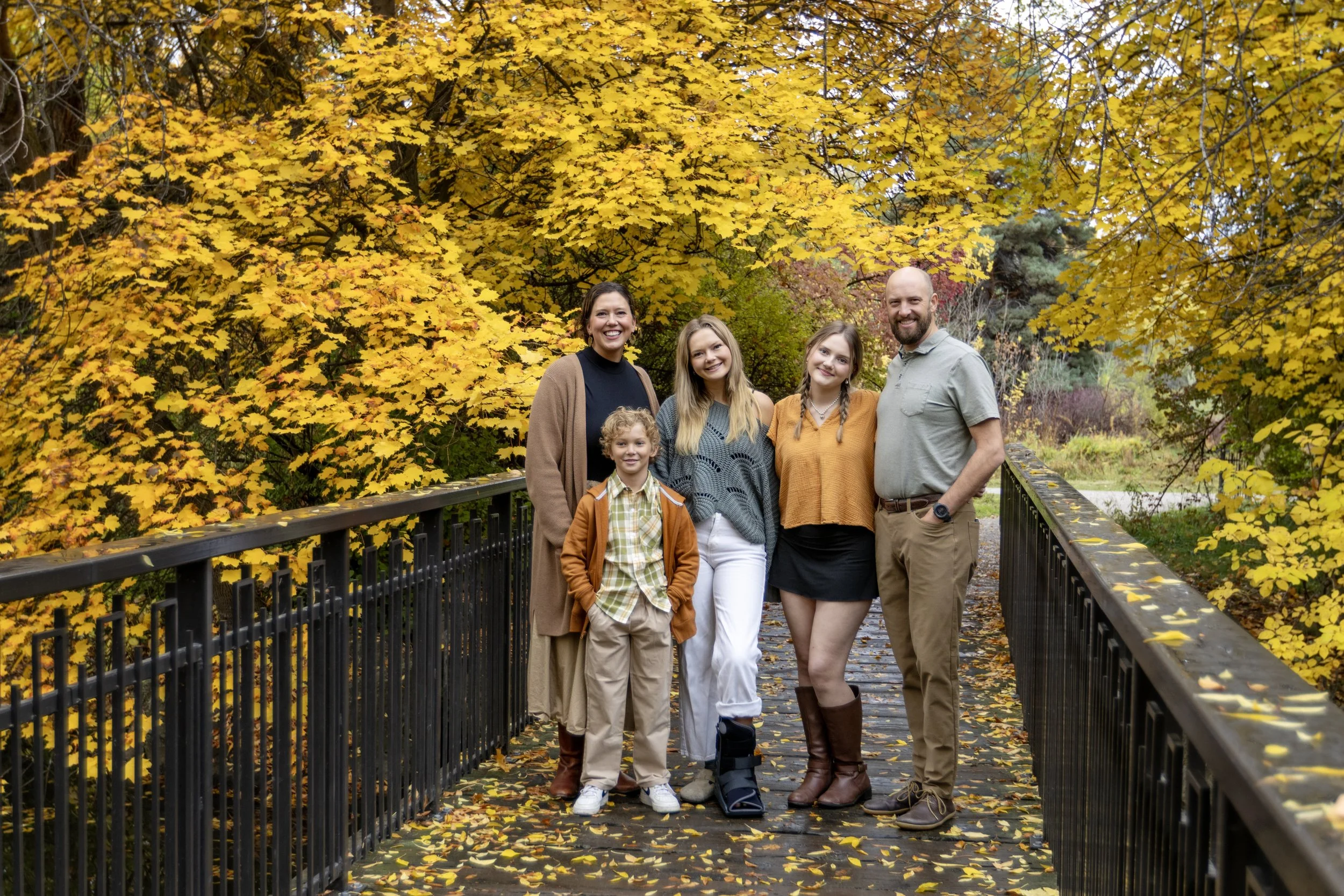 A family of five stands on a wooden bridge with yellow fall foliage behind them. The family includes two women, two men, and a young boy, all smiling at the camera.