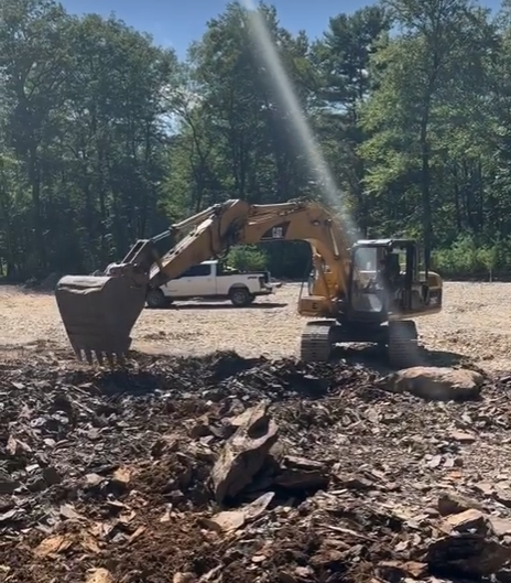 Excavator operating at a construction site with a white pickup truck in the background, surrounded by trees.