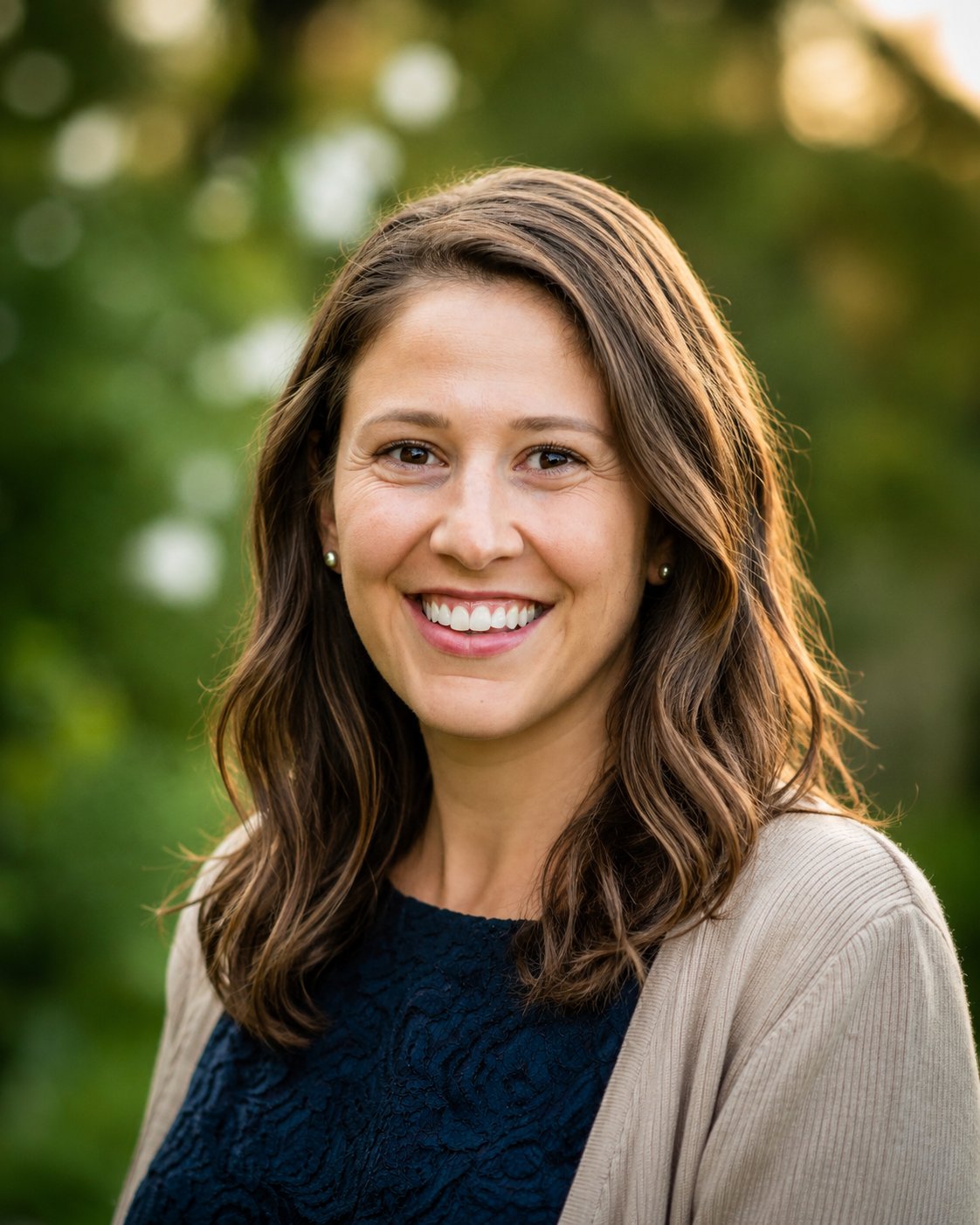 A smiling psychologist with shoulder-length brown hair wearing a navy blue top and beige jacket outdoors during golden hour.