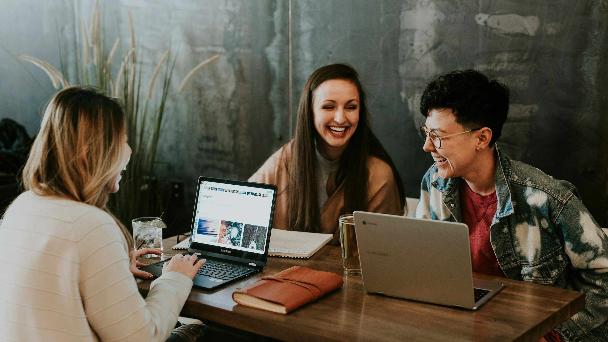 Three women sitting at a table with laptops, laughing and enjoying each other's company in a casual setting.
